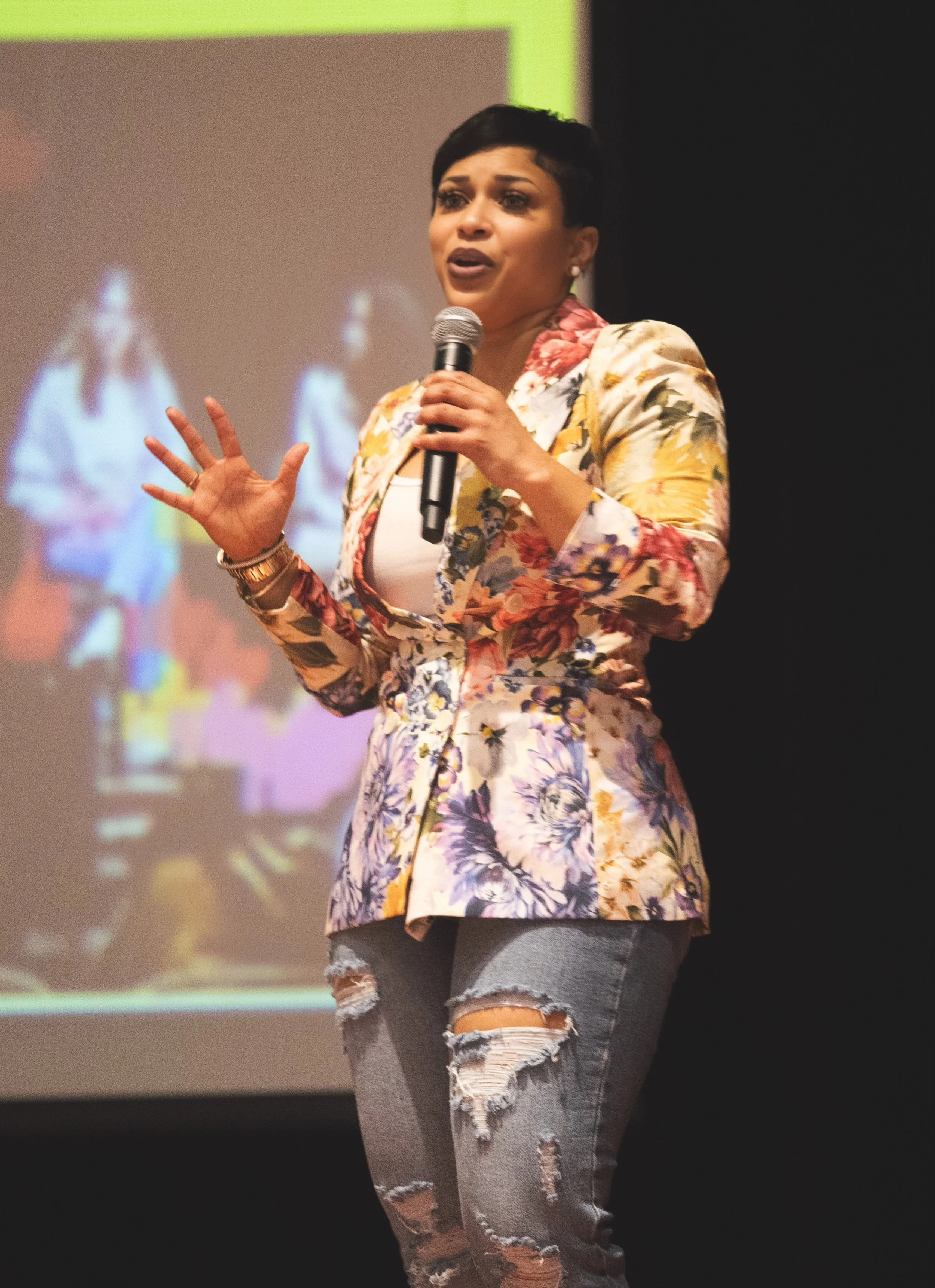 Woman with short dark hair wearing a floral blazer and ripped jeans, holding a microphone and speaking on stage.