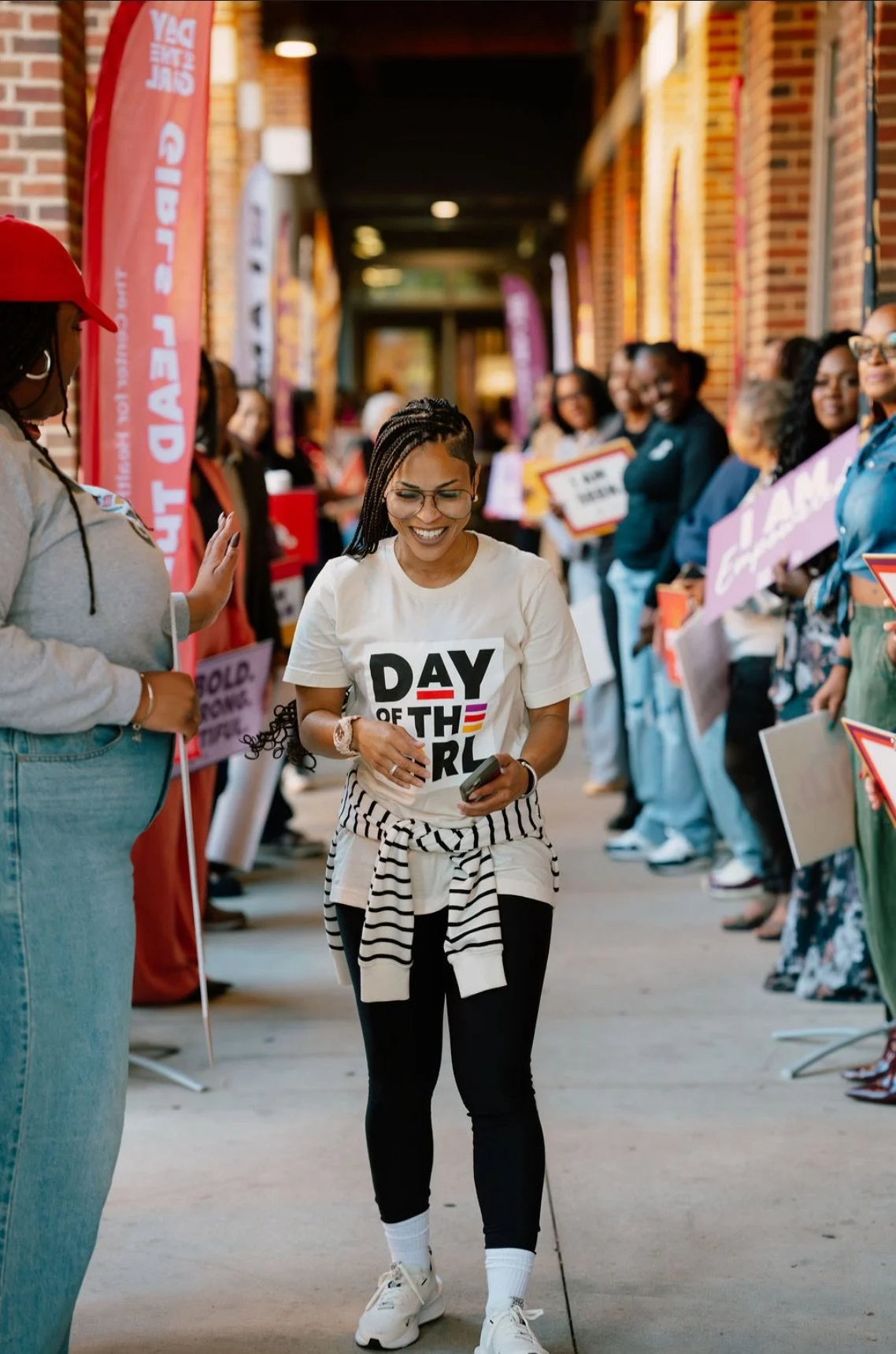 A woman is walking and smiling while holding a phone, wearing a white t-shirt with 'DAY OF THE GIRL' printed on it, surrounded by people holding signs and banners at a gathering or rally celebrating the Day of the Girl.