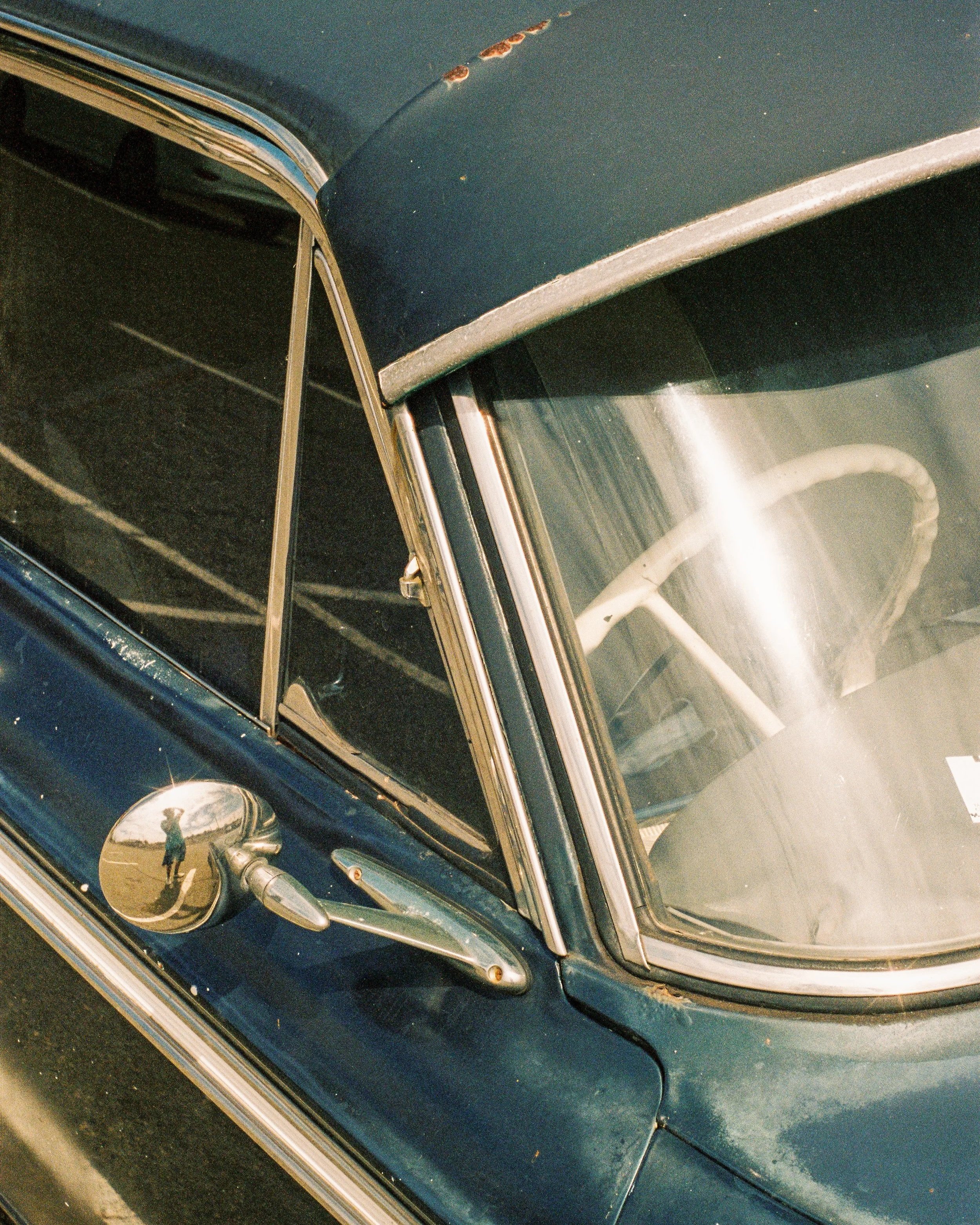 A retro vintage analogue 35mm film picture image of Close-up of a dark blue vintage car with rust spots near the window and chrome details, including the side mirror reflecting the person taking the photo.