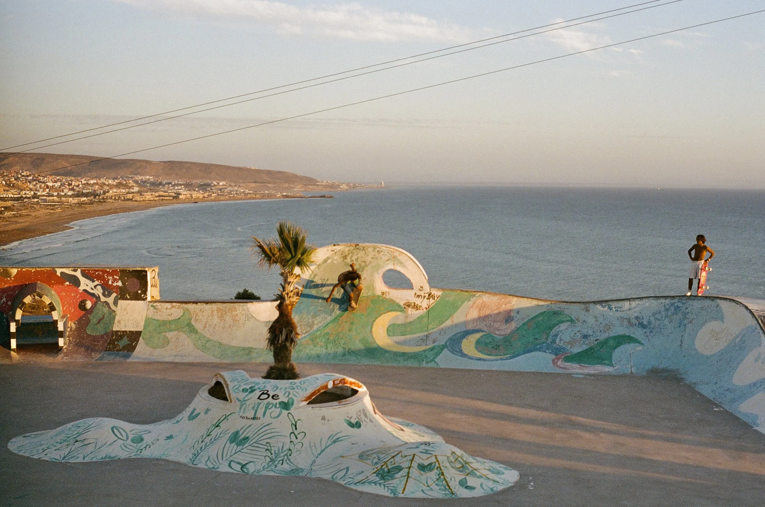 A retro vintage analogue 35mm film picture image of Children playing on a colorful skate park with a mural of waves and abstract designs, overlooking the ocean with a town in the background.