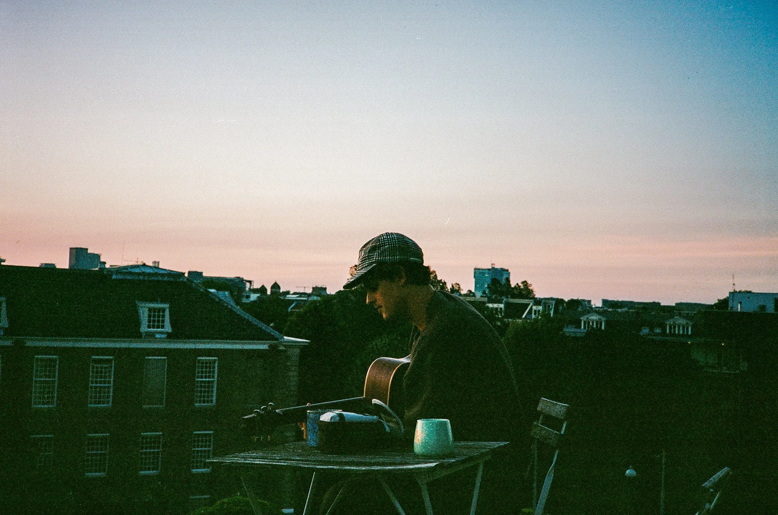 A retro vintage analogue 35mm film picture image of a person wearing a checkered cap and a dark shirt, seen from behind, sitting on a rooftop at sunset with a cityscape background, including buildings and trees.