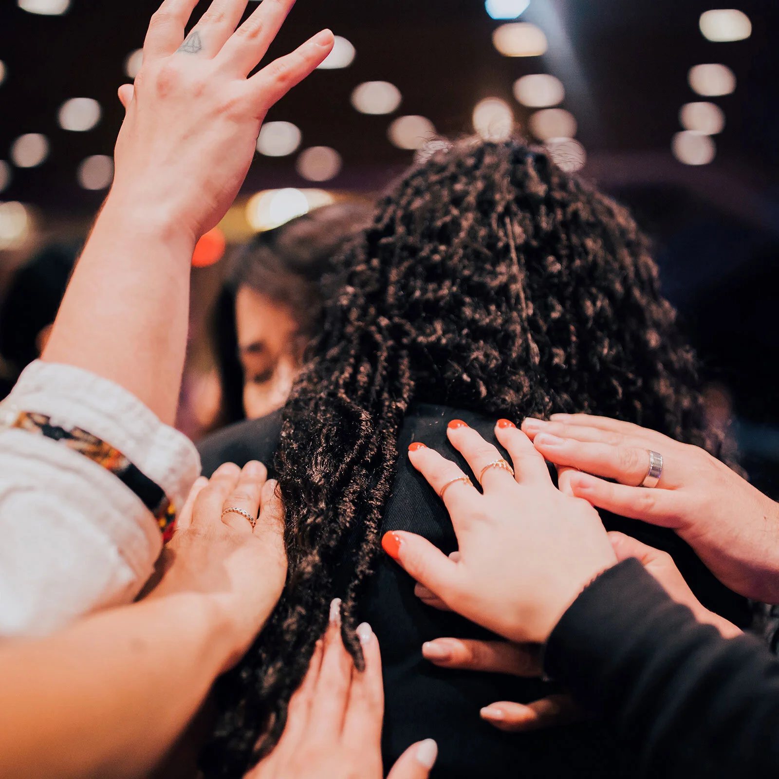 Multiple hands placing hands on a person with curly hair during a group prayer or support moment in a dimly lit setting.