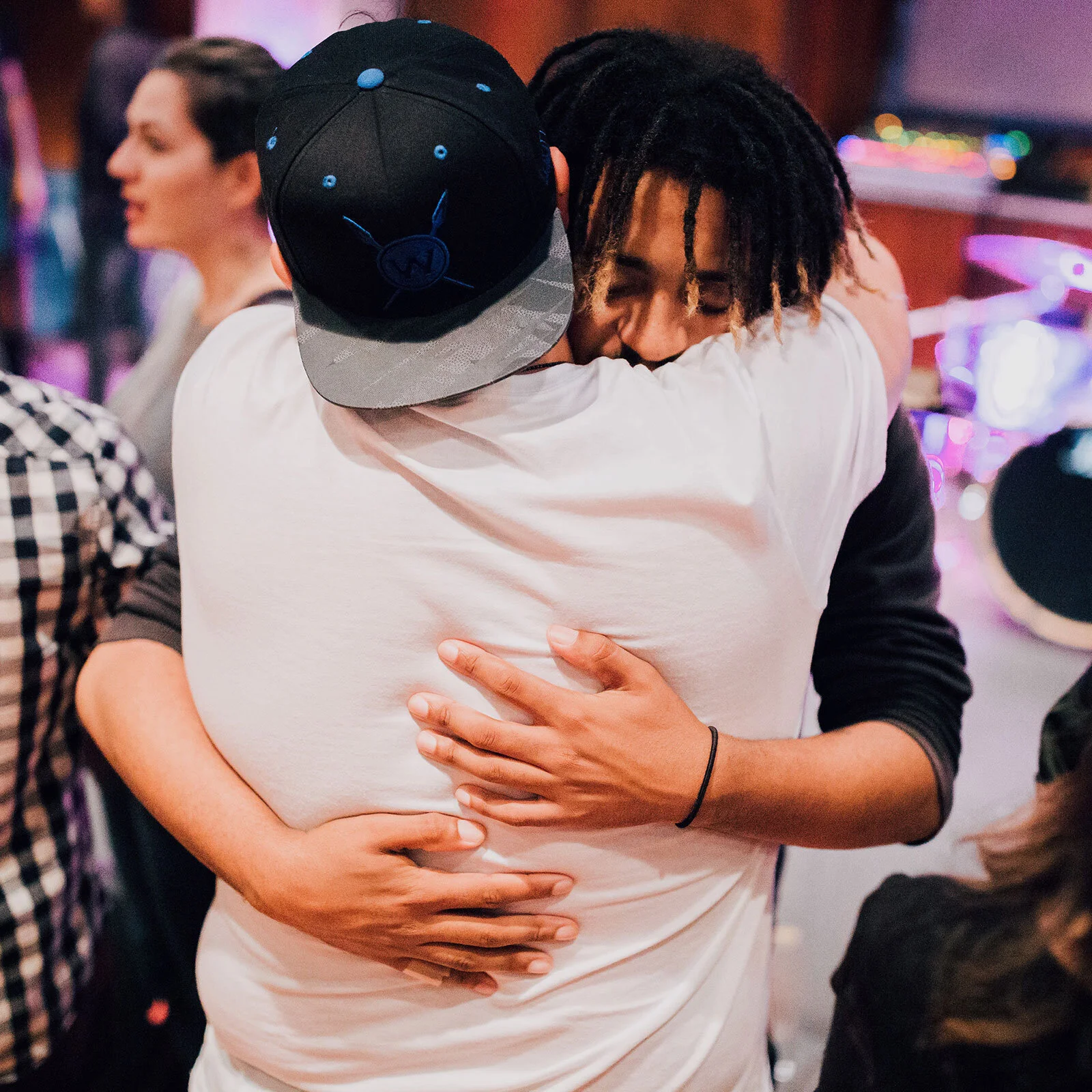 Two people hugging each other warmly in a crowded indoor setting, with one person wearing a black cap and the other with dreadlocks.