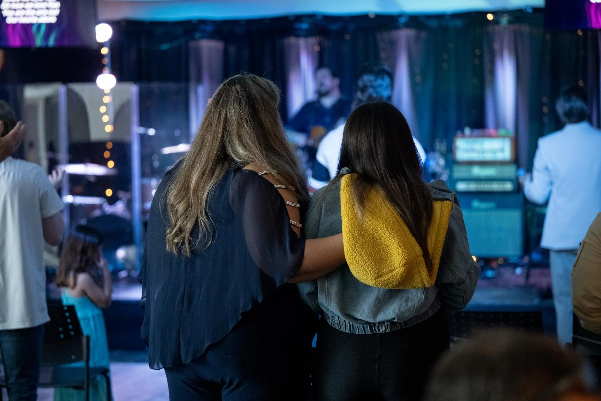 View of a concert or event with people standing and watching a performer on stage, with a dark background and colorful lighting.
