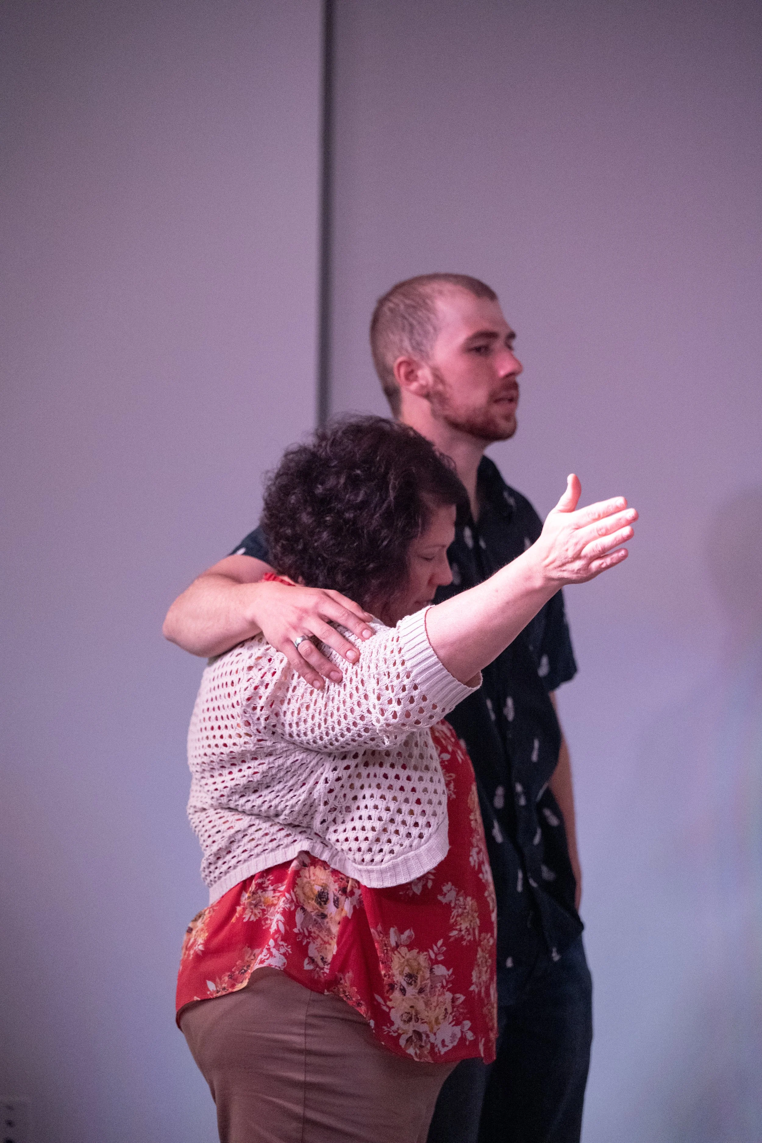 A woman with curly hair embracing a man with short blond hair, both with their eyes closed, in a moment of comfort and support.