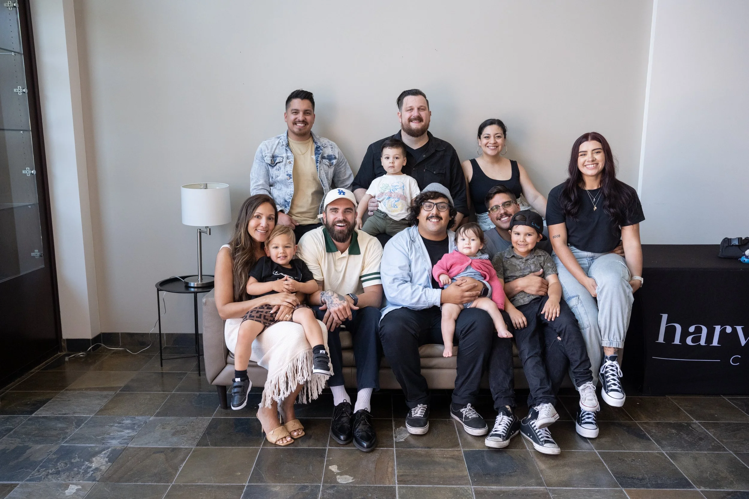A group of twelve diverse people, including children and adults, posed together on a beige sofa against a plain wall in a modern indoor setting.