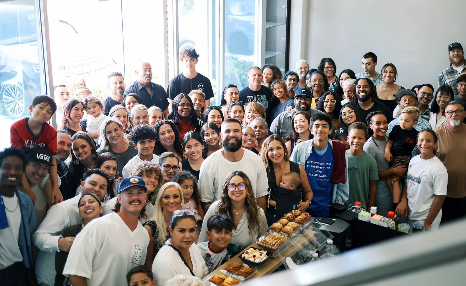 A large group of diverse people of all ages gathered indoors for a celebration or event, with snacks and desserts on a table in the foreground.