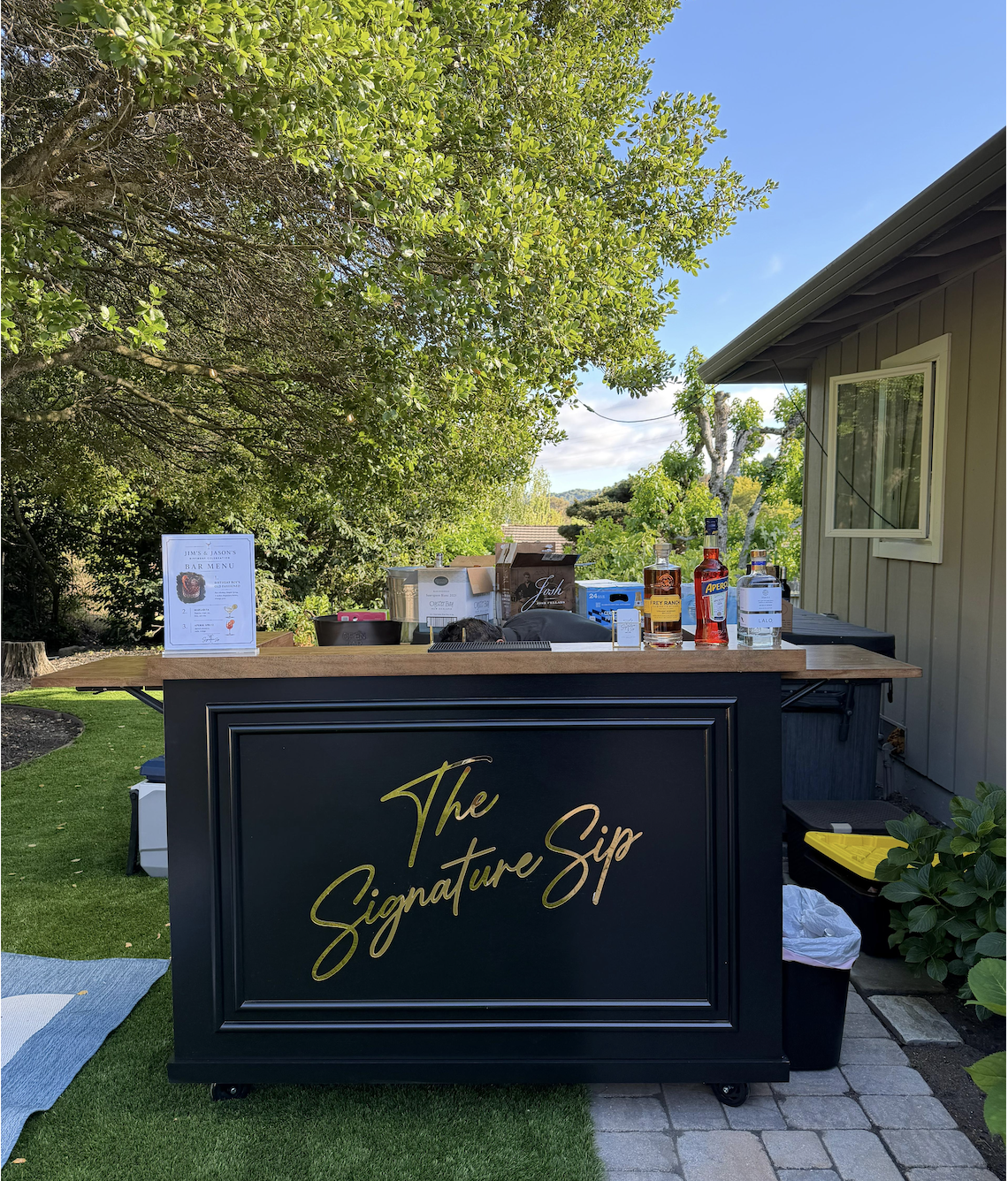 An outdoor bar setup with a black and gold sign that says 'The Signature Sip' on a wooden counter, with bottles of alcohol, cups, and other bar items, surrounded by green trees and a house on the right side, under a clear blue sky.