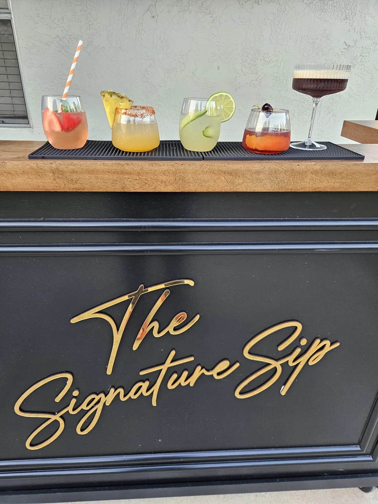 Six colorful cocktails on a bar counter, with a black sign with gold cursive writing in the foreground.