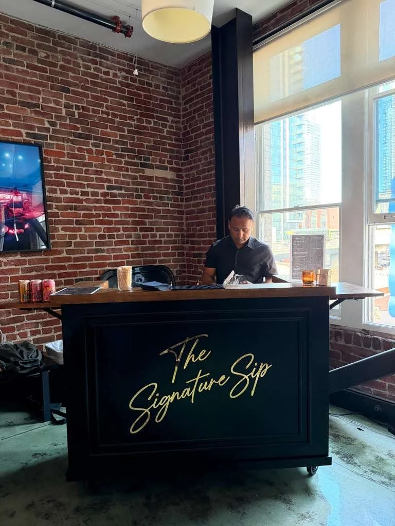 A man working at a black bar with gold lettering that reads 'The Signature Sip,' in front of large windows and exposed brick walls in a modern setting.