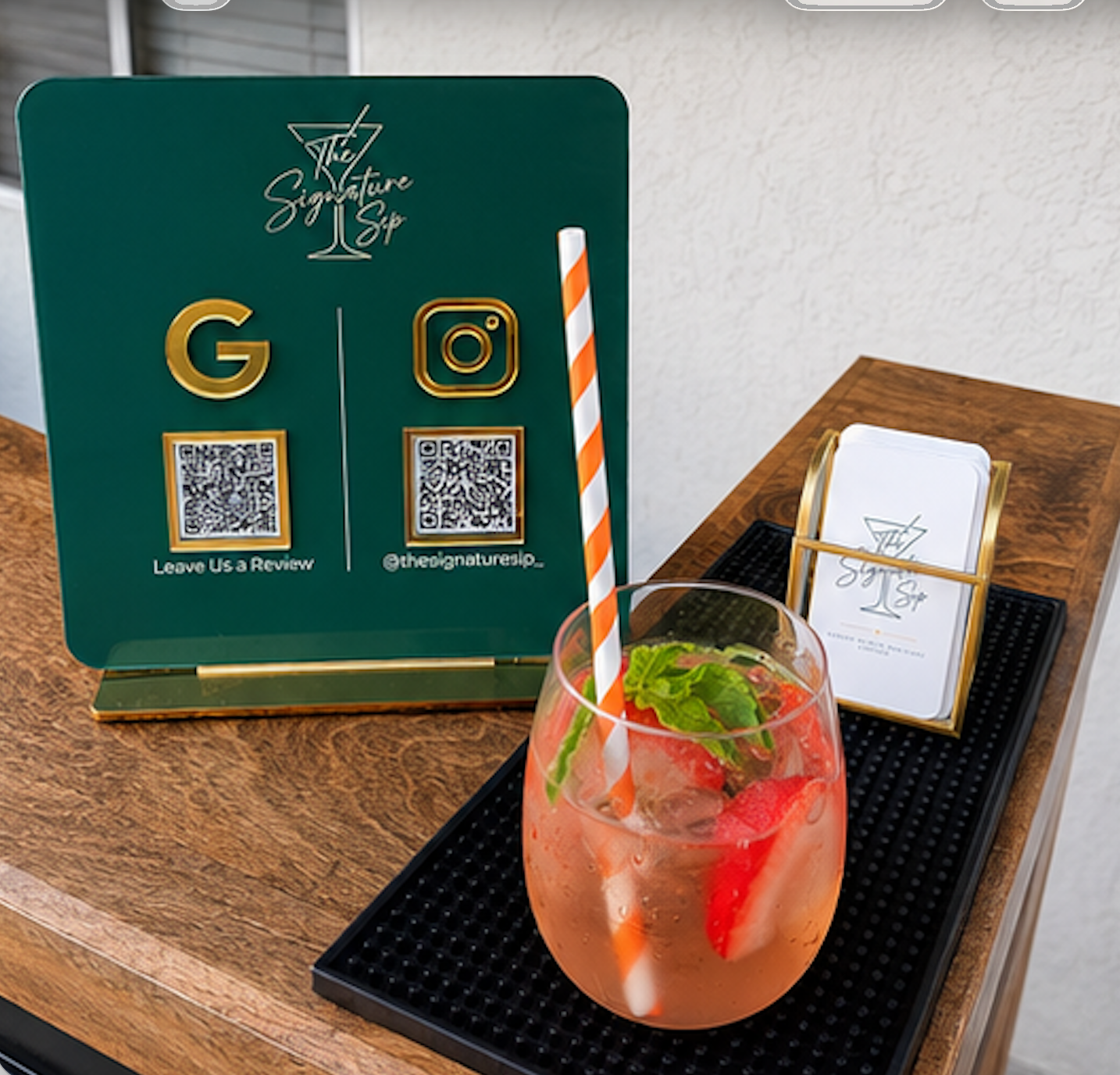 A wooden table with a dark tray holding a pink cocktail with strawberries, mint, and a striped straw. A green tabletop sign with QR codes and social media info, along with a menu holder next to it.