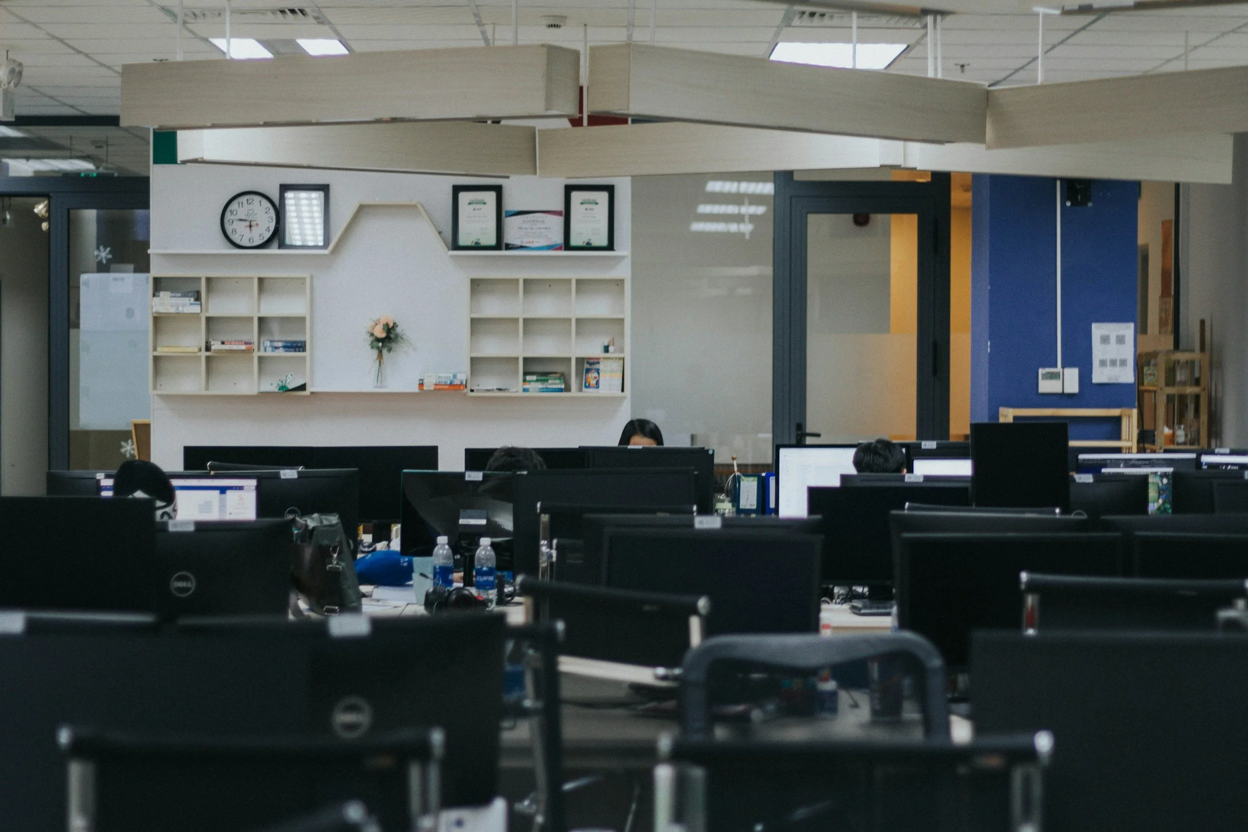 Office workspace with multiple computer desks, some water bottles, and a white wall with a clock, framed awards, and a shelf with books, in a modern office setting.