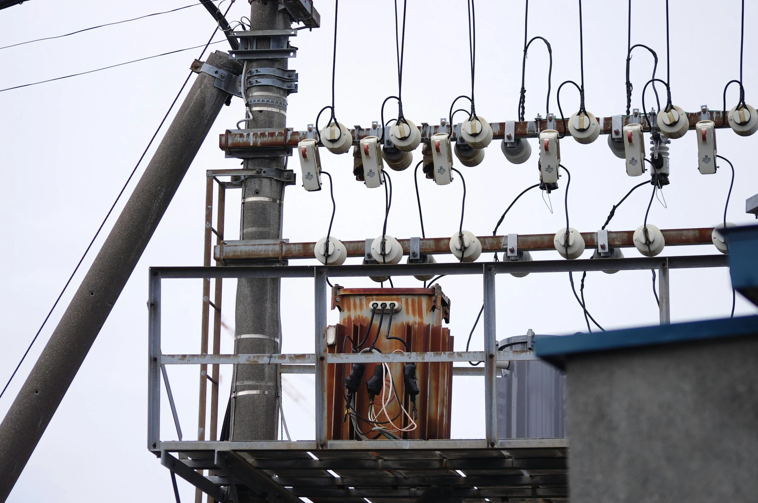 Close-up of an electrical utility pole with insulators, wires, and electrical equipment mounted on a metal platform against a cloudy sky.