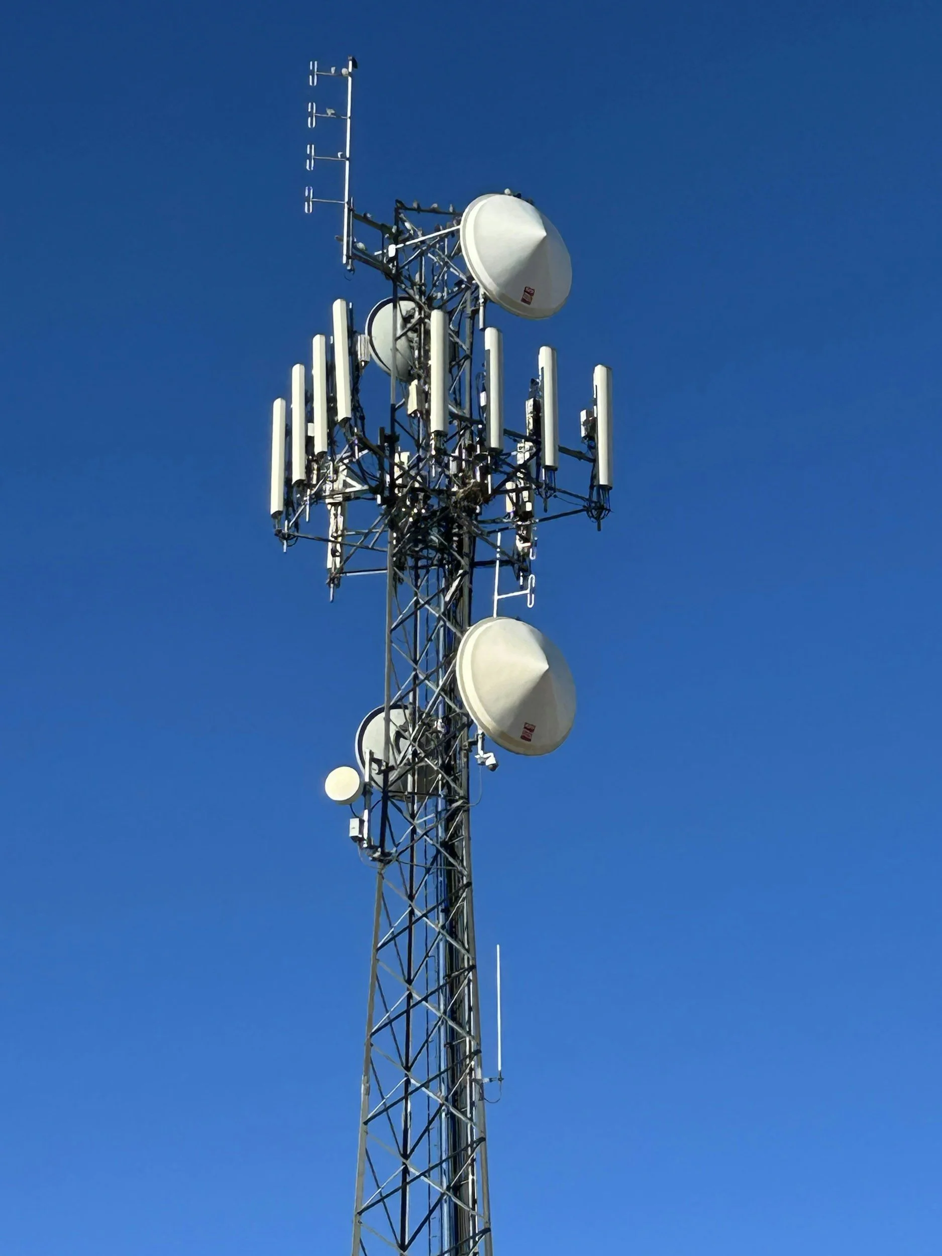 A tall communication tower with multiple antennas and satellite dishes against a clear blue sky.