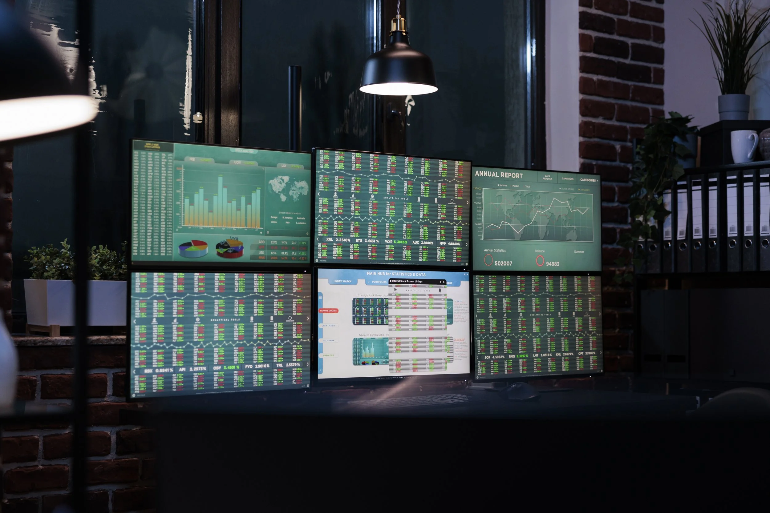 Multiple computer monitors displaying financial data, charts, and graphs, set on a desk against a brick wall with decorative plants and a hanging lamp.