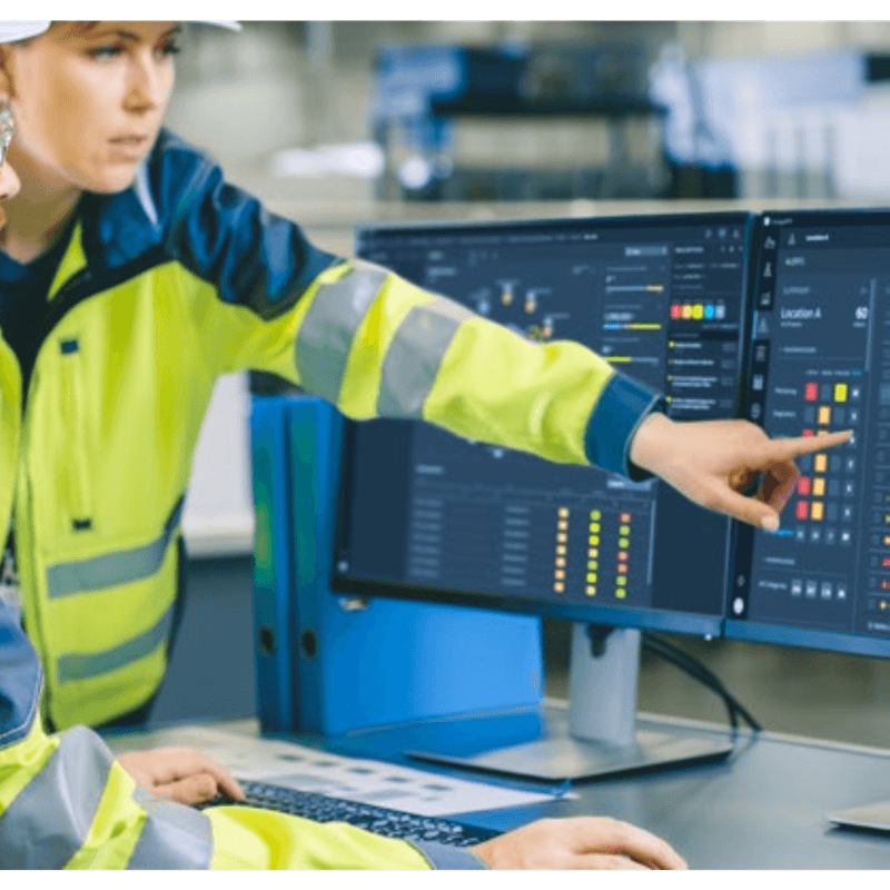 Person wearing a high-visibility safety vest pointing at computer screens with data charts and graphs.