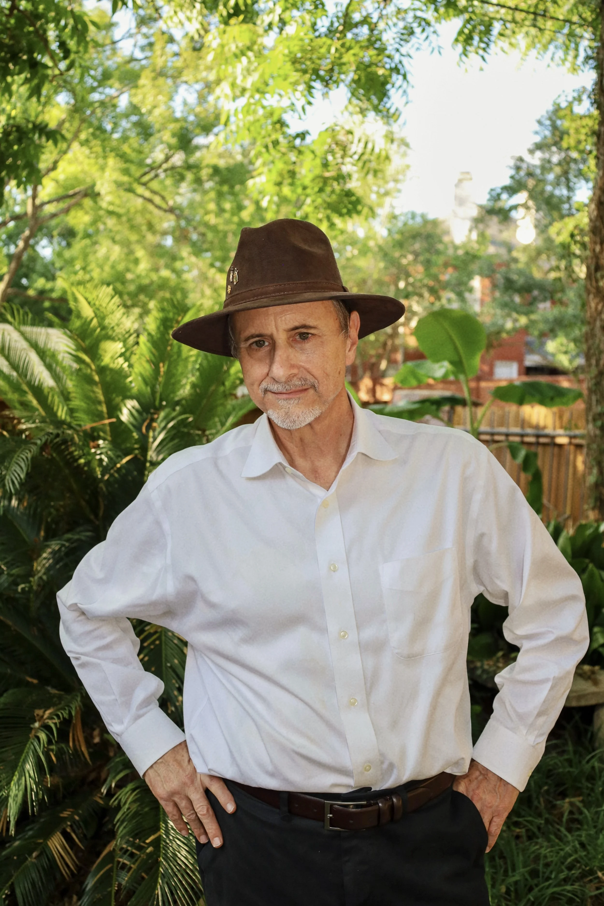 Charles Stewart wearing a white shirt and a brown wide-brimmed hat, standing outdoors in a lush green garden with trees and plants, posing with hands on hips.