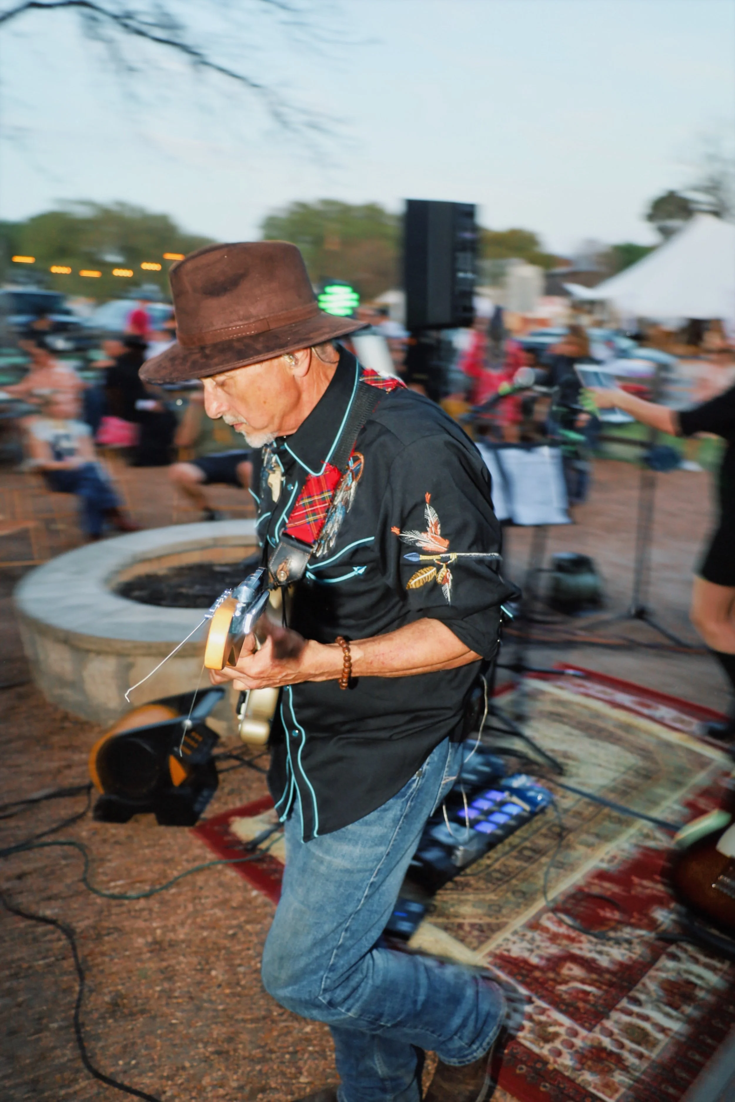 Charles Stewart wearing a brown hat and black shirt with floral embroidery plays an electric guitar at an outdoor music event. He is kneeling on a patterned rug surrounded by musical equipment, with a crowd and tents visible in the background.