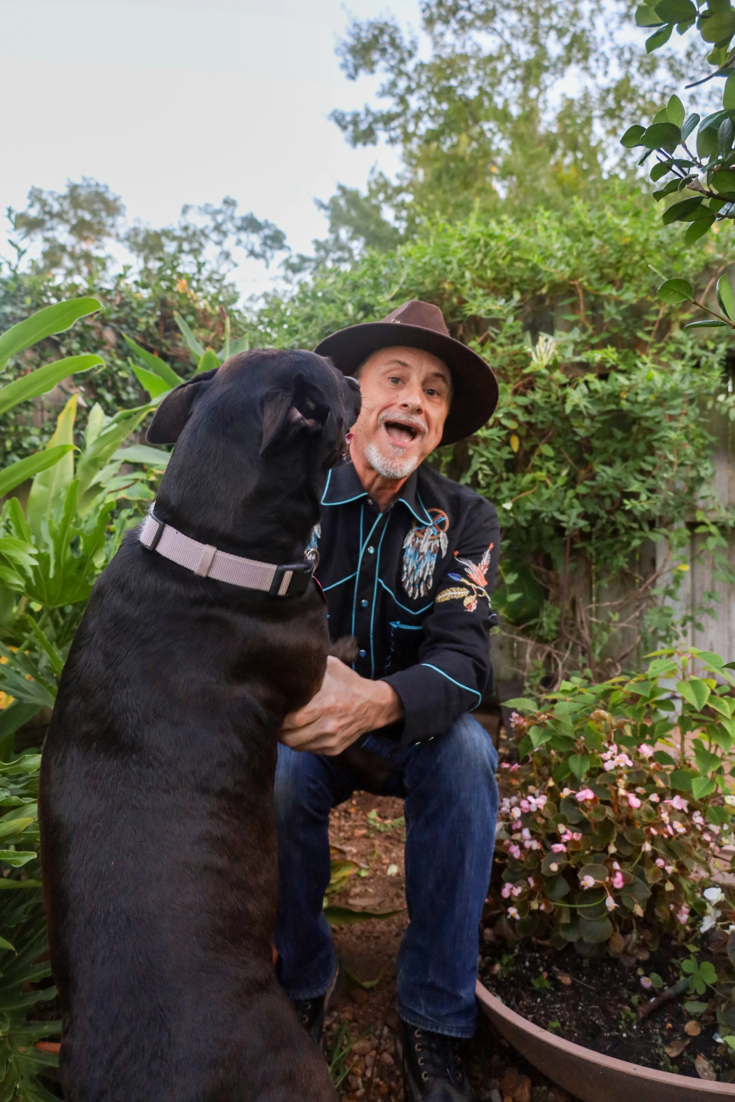 Charles Stewart wearing a wide-brimmed brown hat and a black shirt with colorful floral embroidery is sitting outdoors in a garden, holding a black dog with a light-colored collar; they are surrounded by lush green plants and flowers..