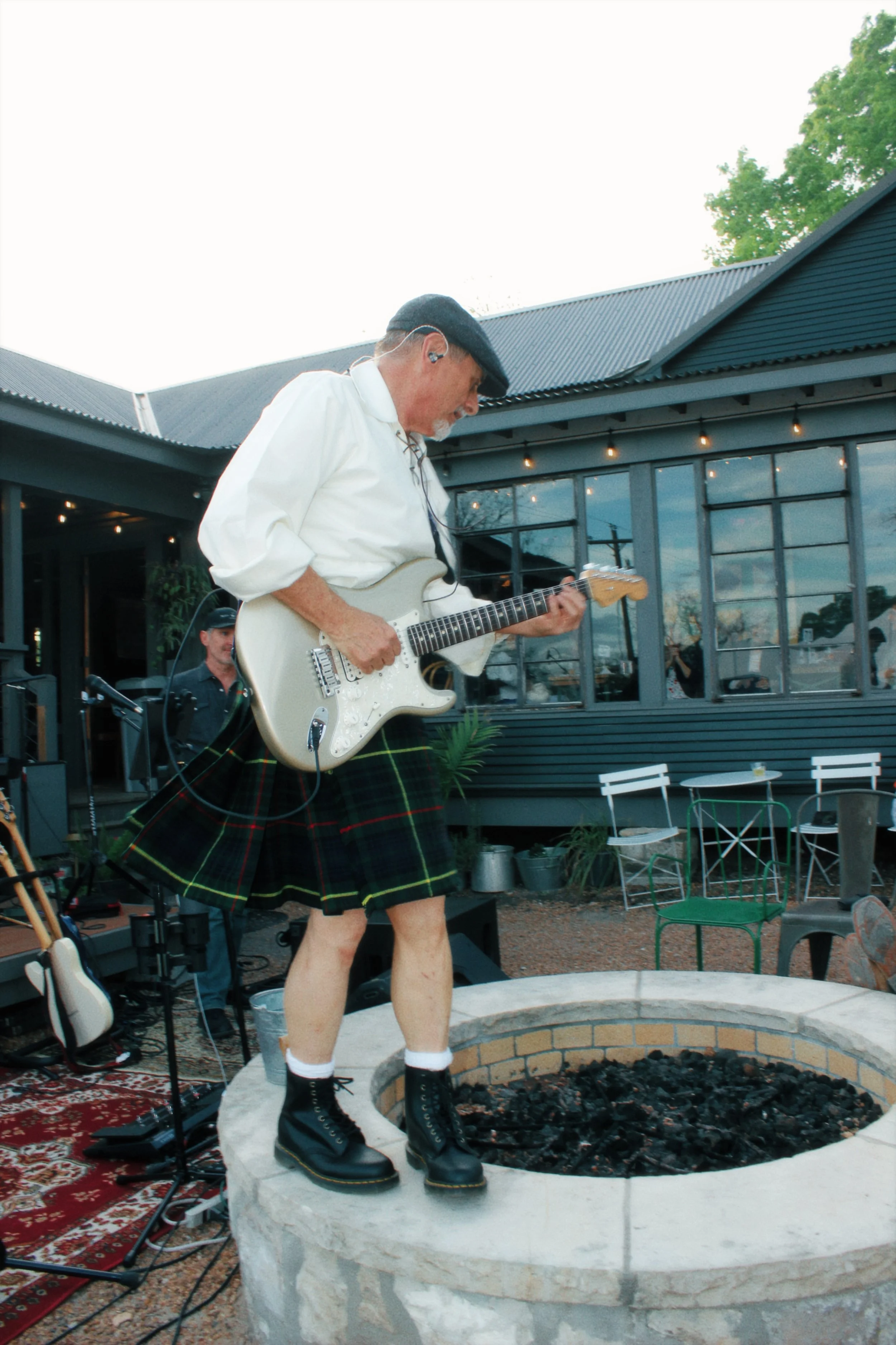 Charles Stewart wearing a white shirt, a black cap, a Stewart Plaid tartan kilt, and black boots is playing an electric guitar outdoors during daytime. He is standing on a stone platform in front of a fire pit and is surrounded by musical equipment.