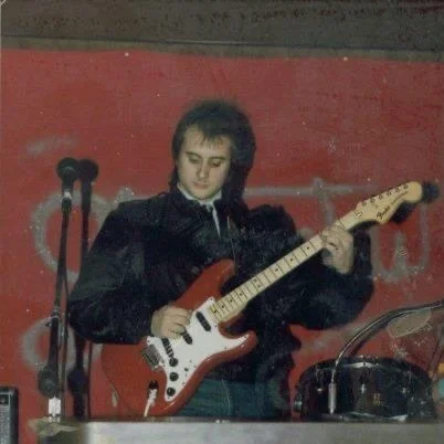 Charles Stewart playing a red stratocaster guitar on stage with a microphone in front of them in the 1980s.