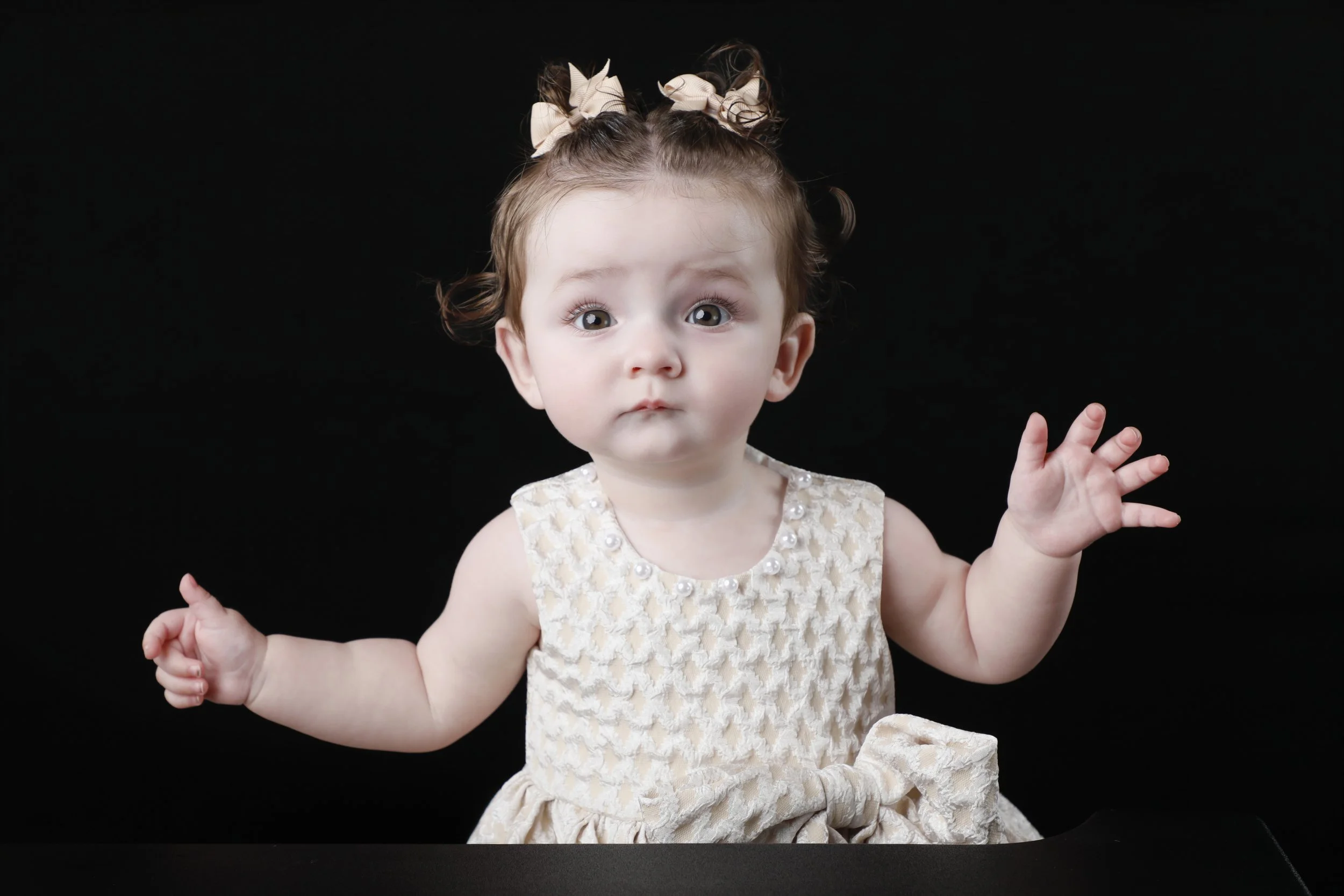 A young girl with curly light brown hair styled in two buns with cream-colored bows, wearing a dress with pearl details, is sitting against a black background with her arms raised and eyes wide open.