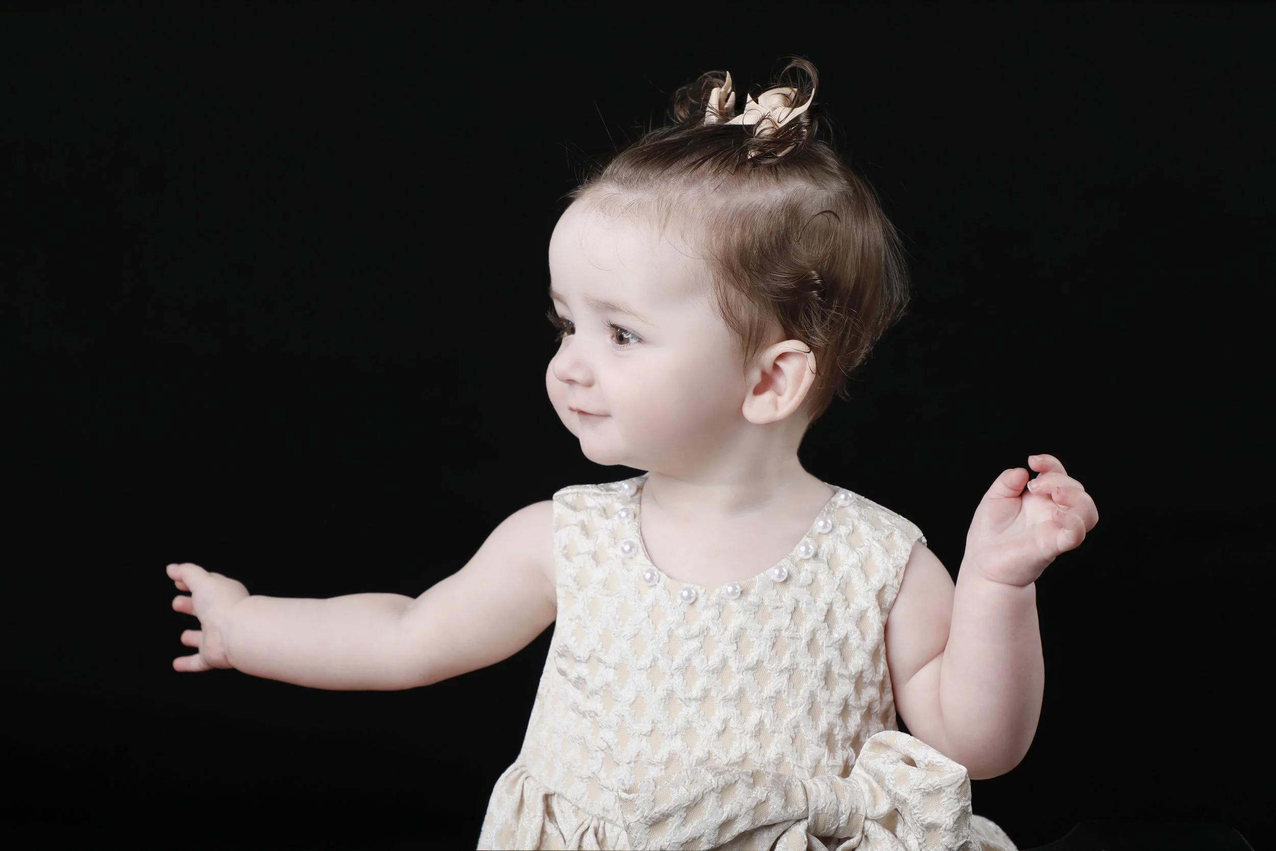 A young girl with curly brown hair in a top bun with a bow, wearing a beige dress with pearl accents, appears to be reaching out with her left hand and looking to her left against a black background.