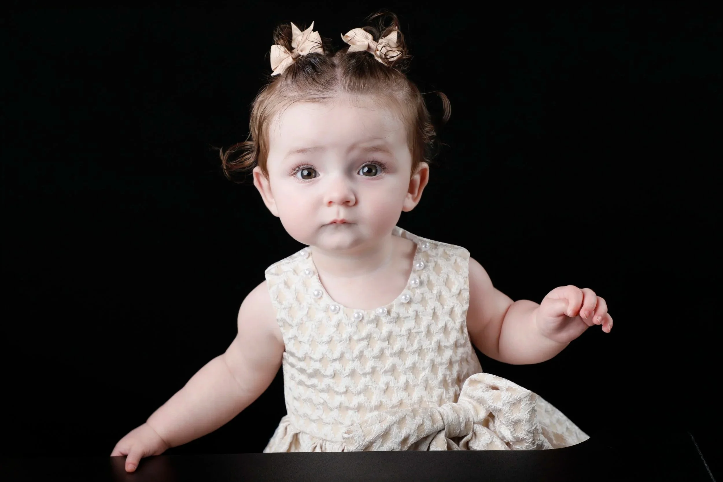 A young girl with curly brown hair tied in pigtails with beige bows, wearing a cream-colored dress with pearl accents, looking directly at the camera against a black background.