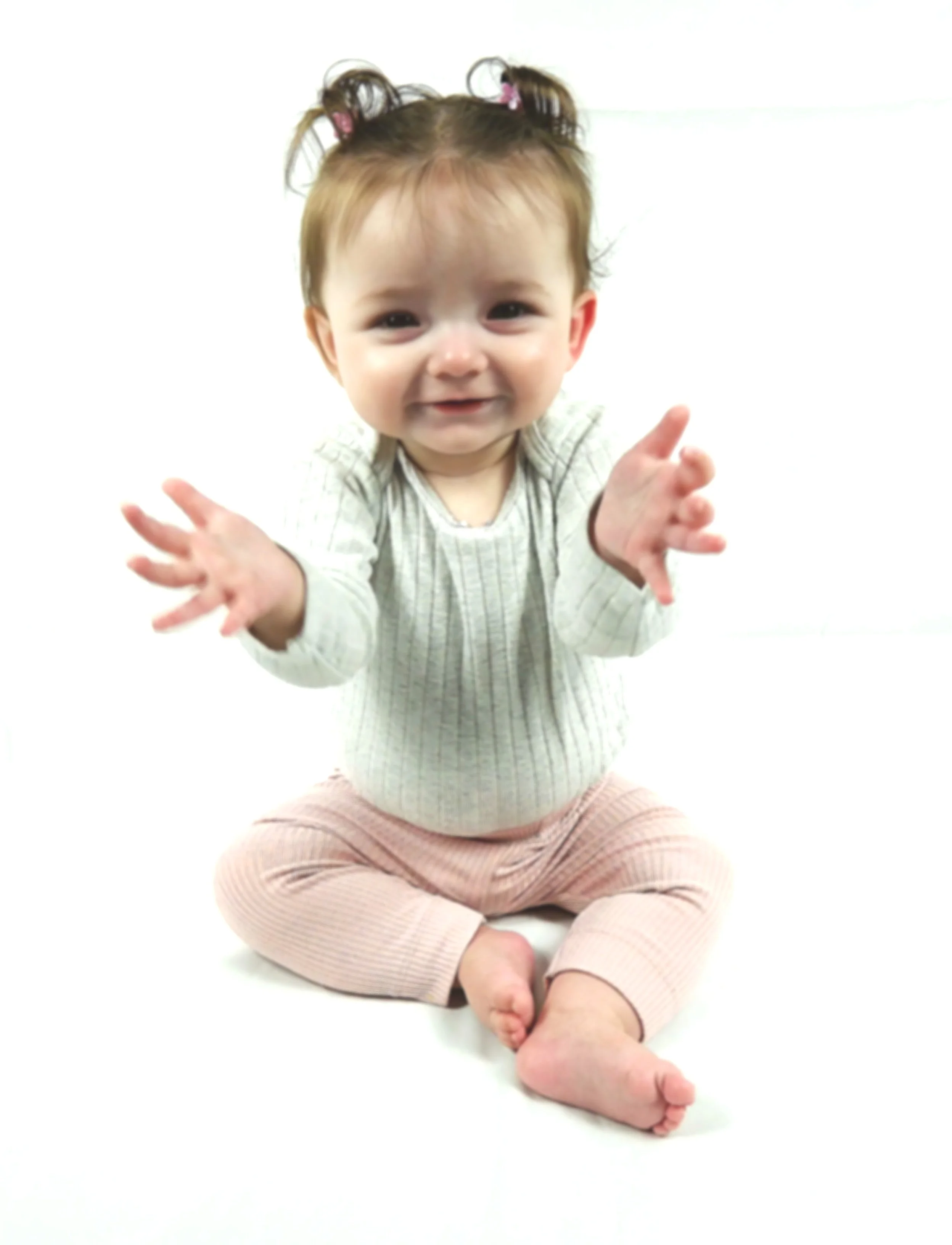 A smiling baby girl with brown hair in pigtails, wearing a beige striped long sleeve shirt and pink pants, sitting on the floor with arms outstretched.