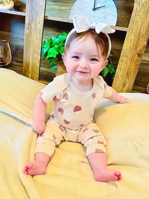 A smiling baby girl with a white bow headband, sitting on a yellow bed wearing cream-colored pajamas with a red mushroom print in a cozy room with wooden decor, a clock, and plants in the background.