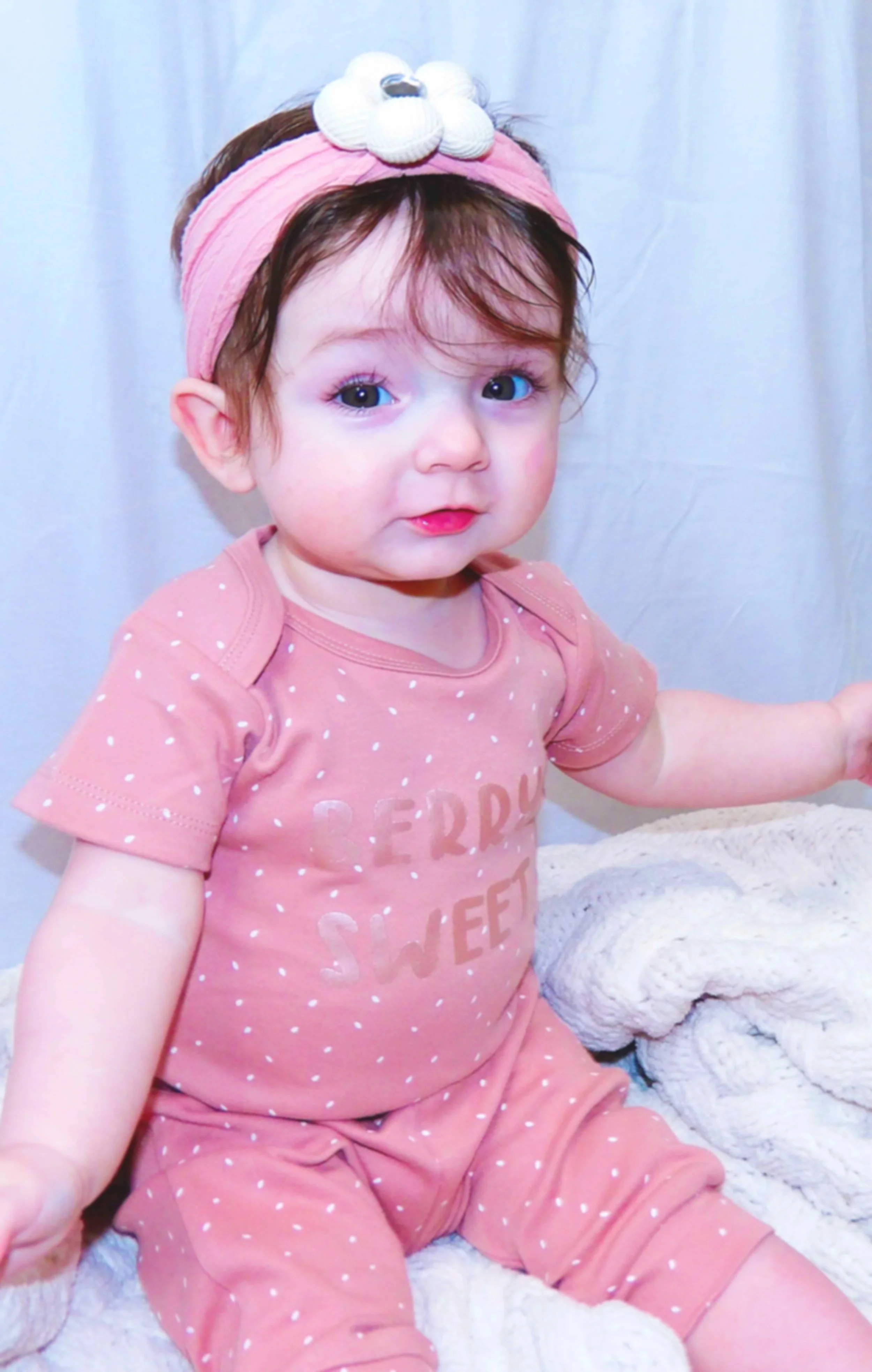 A young girl with light brown hair, blue eyes, and fair skin wearing a pink polka-dotted outfit and a pink headband with a white flower. She is looking at the camera and sitting on a soft white surface with a plain light background.
