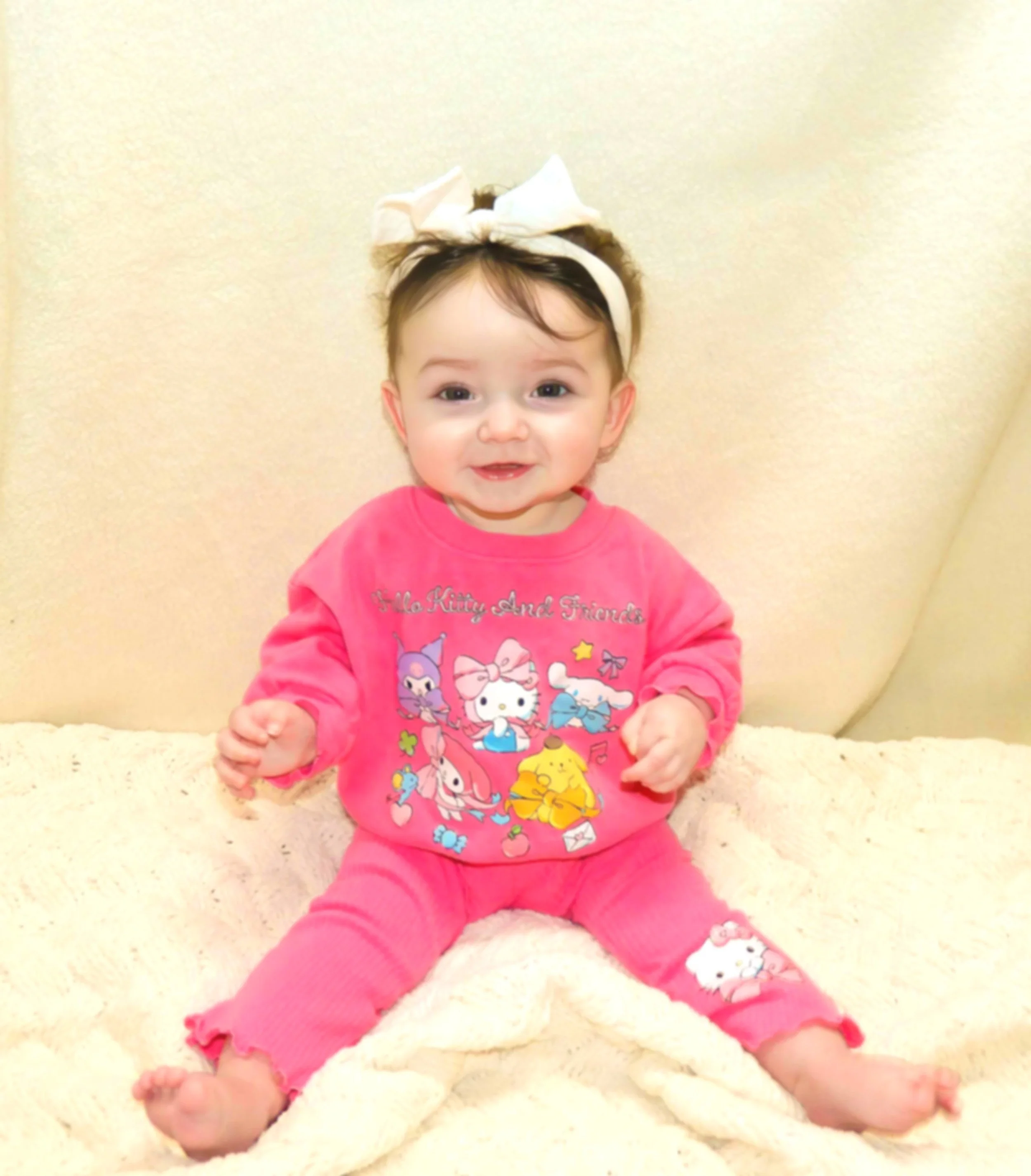 Smiling baby girl sitting on a fluffy cream-colored blanket, wearing pink pajamas with cartoon characters and a large beige headband with a bow.