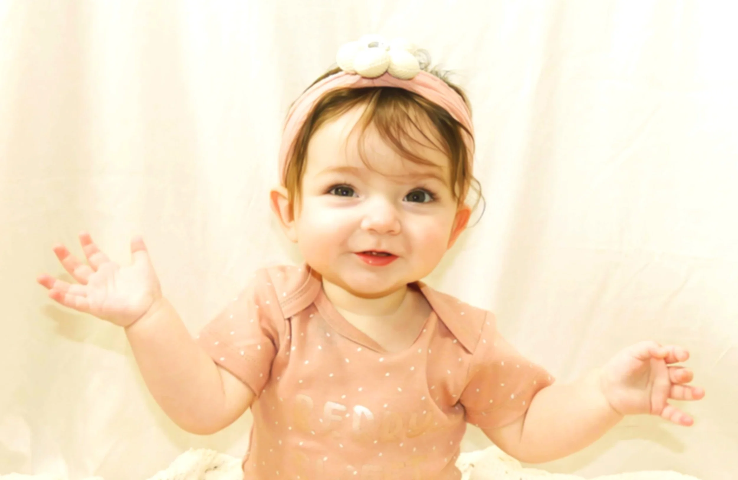 A young girl with red hair, wearing a peach-colored dress with white dots and a matching headband with a bow, sitting against a beige background, waving with her left hand and smiling at the camera.