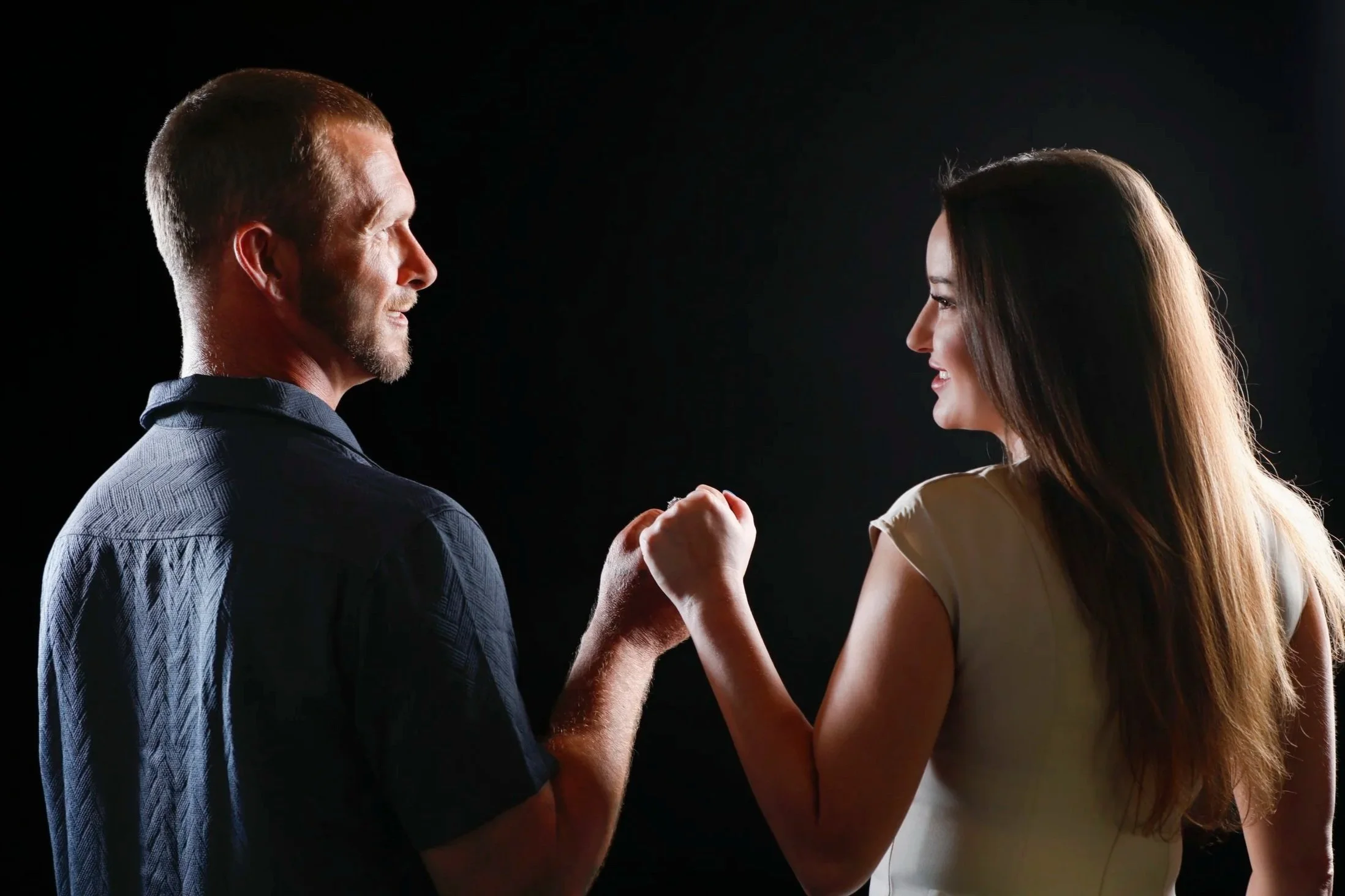 A man and woman face to face, smiling and holding hands against a black background.