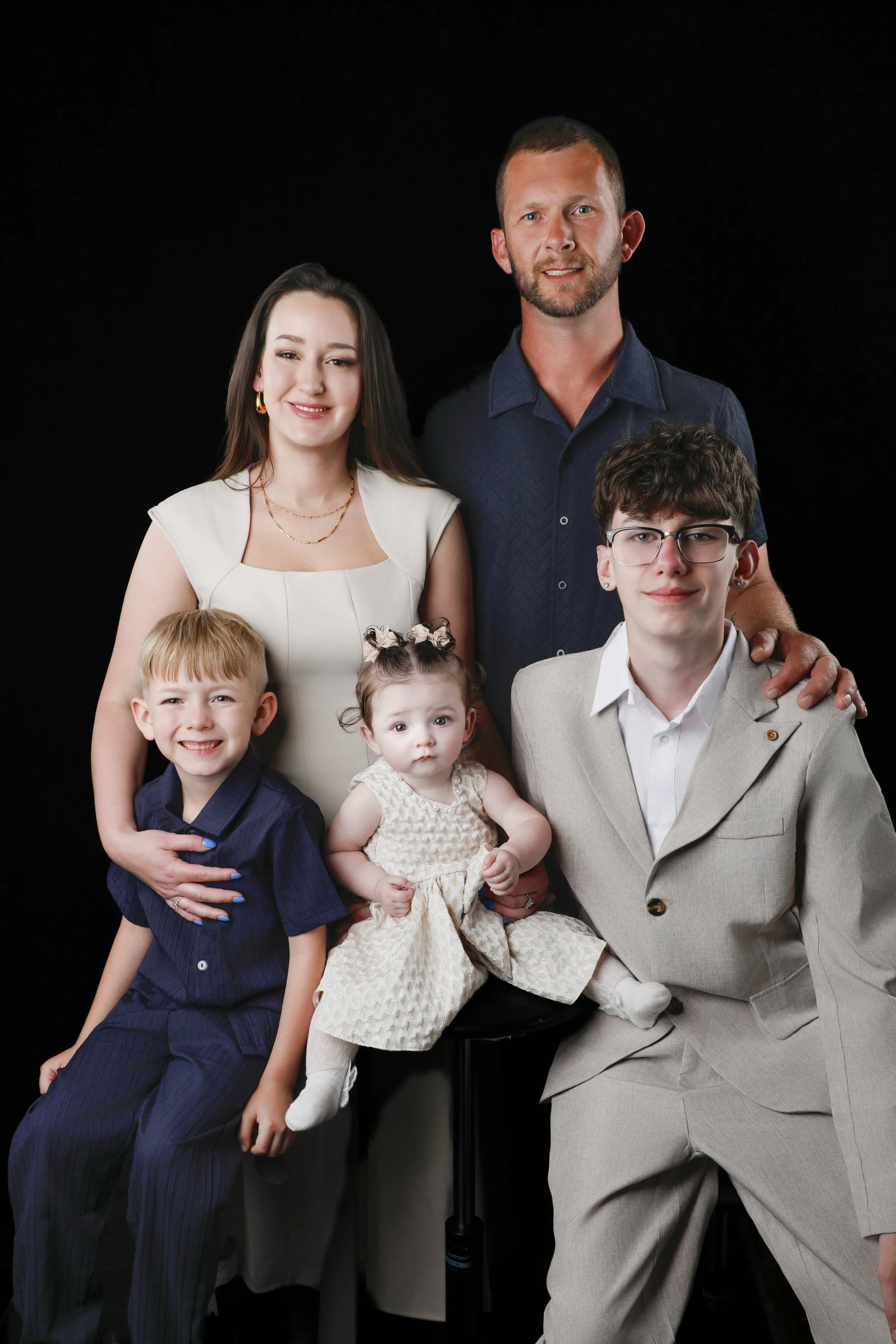 A family portrait with five members against a black background. The woman has long dark hair, is wearing a white sleeveless dress, and gold jewelry. The man has short hair, a beard, and is dressed in a blue shirt. The boy has glasses, short brown hai