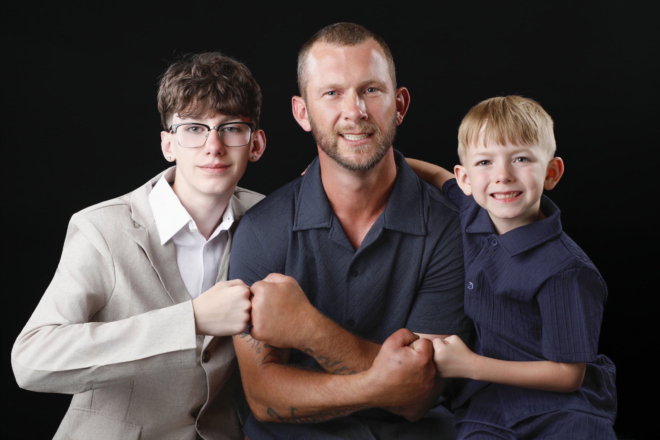 A man and two children posing together, with the children holding the man's hands in a supportive manner, against a black background.