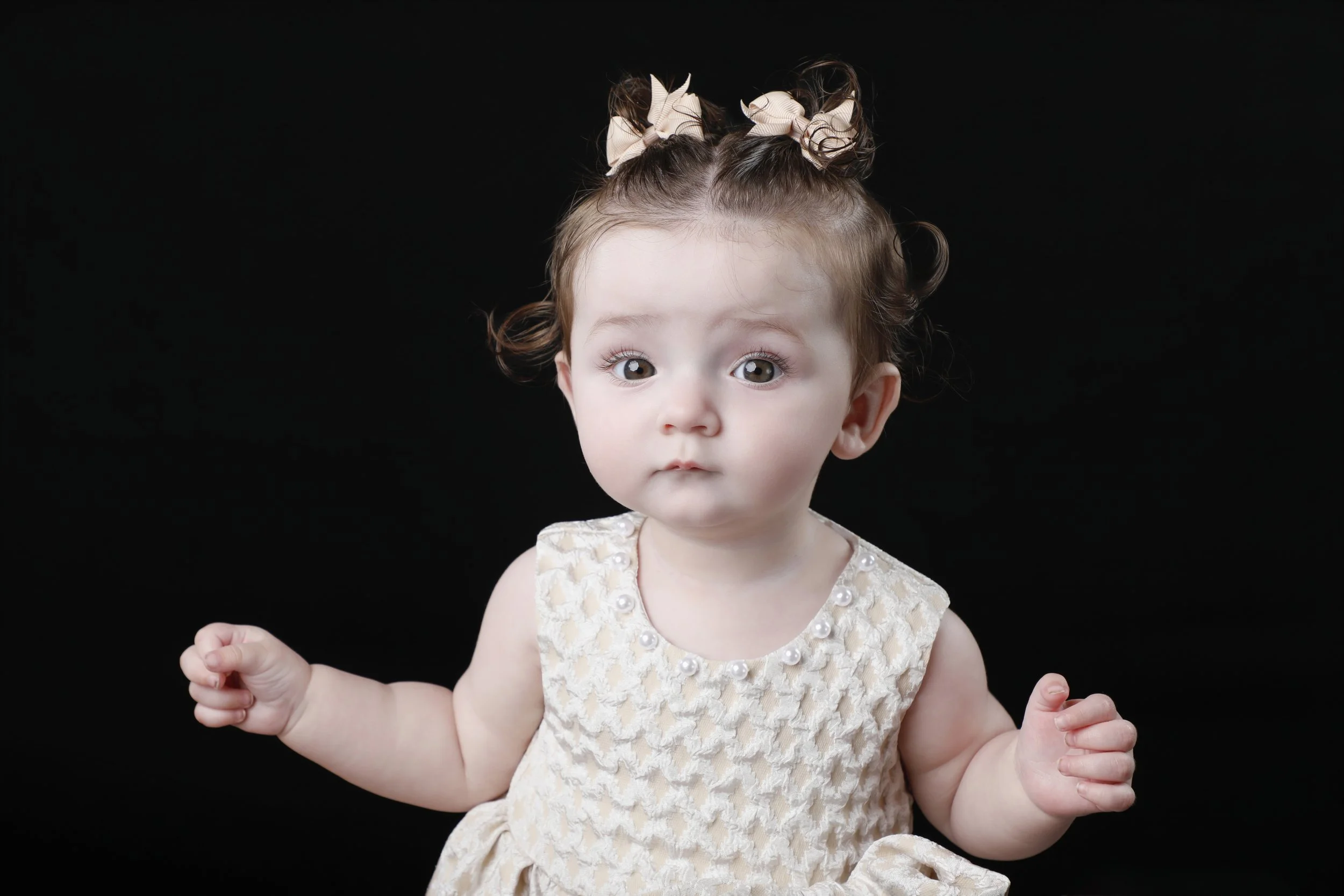 A young girl with curly light brown hair tied in two puffy buns with beige bows, looking surprised with wide eyes, posed against a black background, wearing a cream-colored dress with a textured pattern and pearl embellishments.