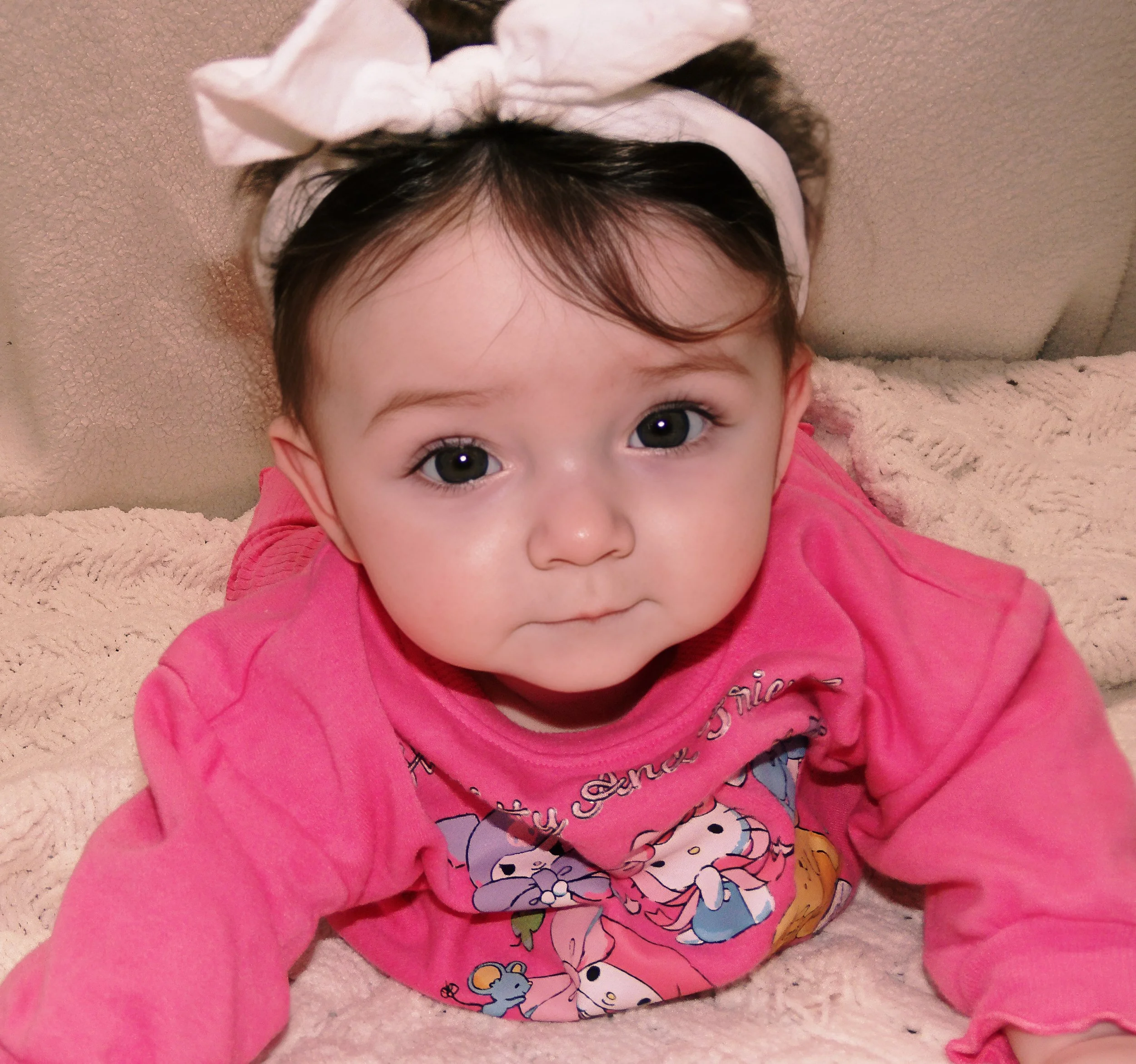 Close-up of a young child's face with big eyes, wearing a pink shirt featuring cartoon characters and a white headband with a bow, lying on a textured beige surface.