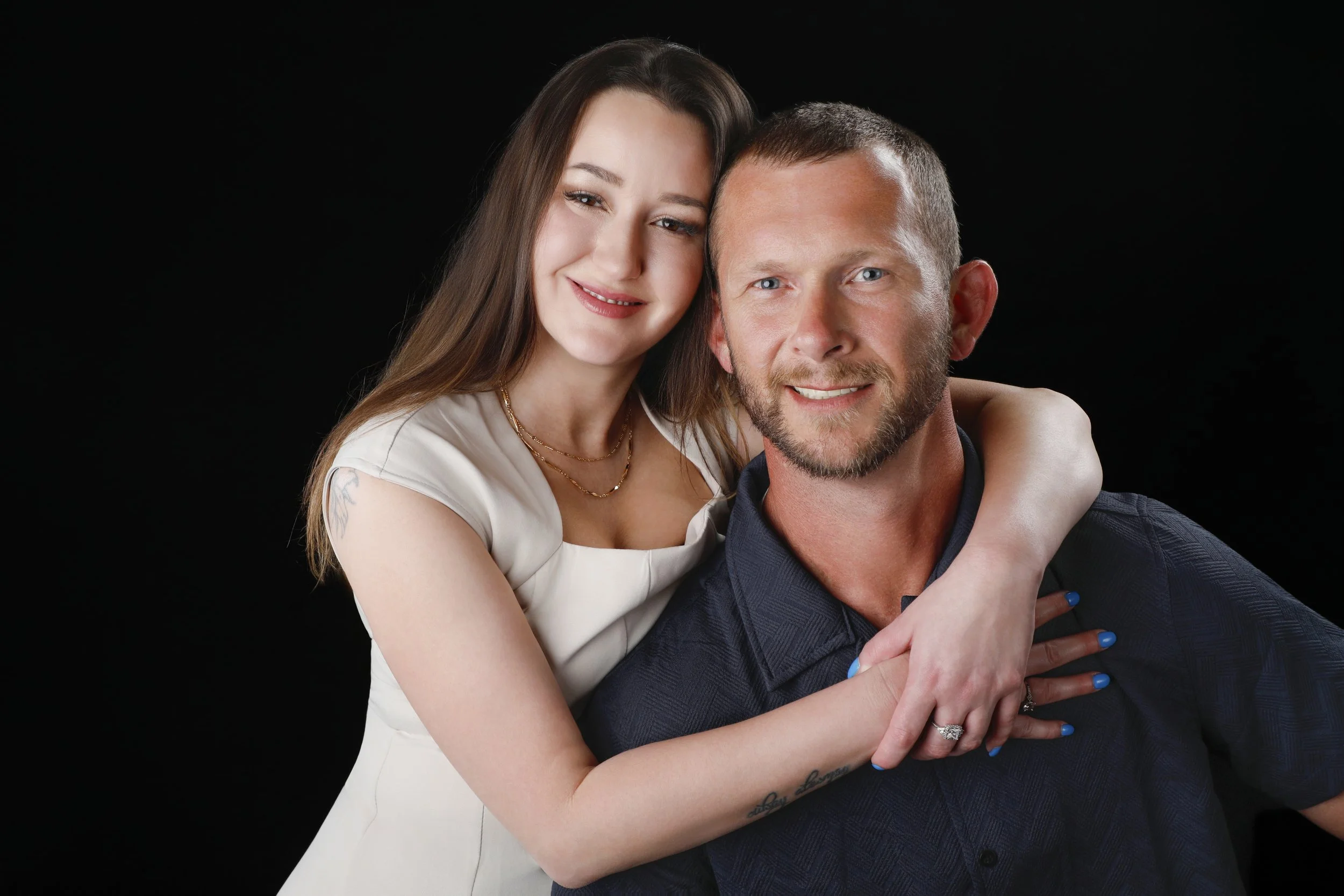 A smiling young woman with long brown hair hugging a smiling man with short hair and a beard against a black background.