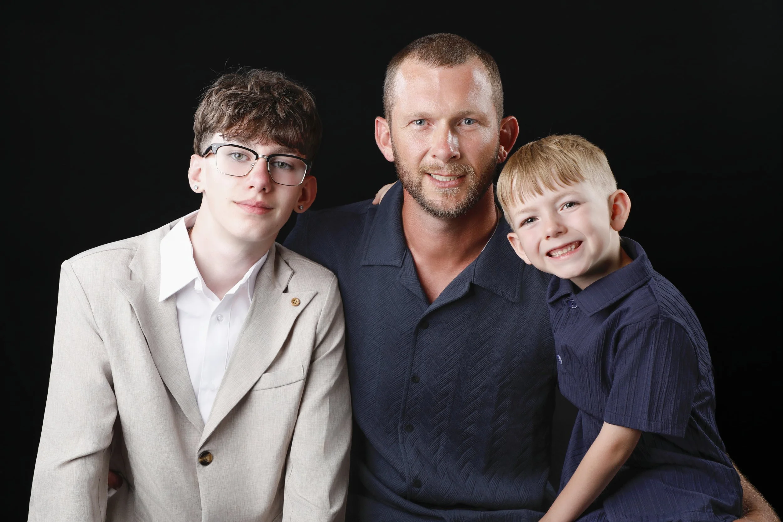 A man with short hair and a beard, wearing a navy blue shirt, is sitting with a boy with blond hair and a dark shirt, smiling. A girl with short hair, glasses, earrings, and a light-colored blazer is sitting beside them against a black background.