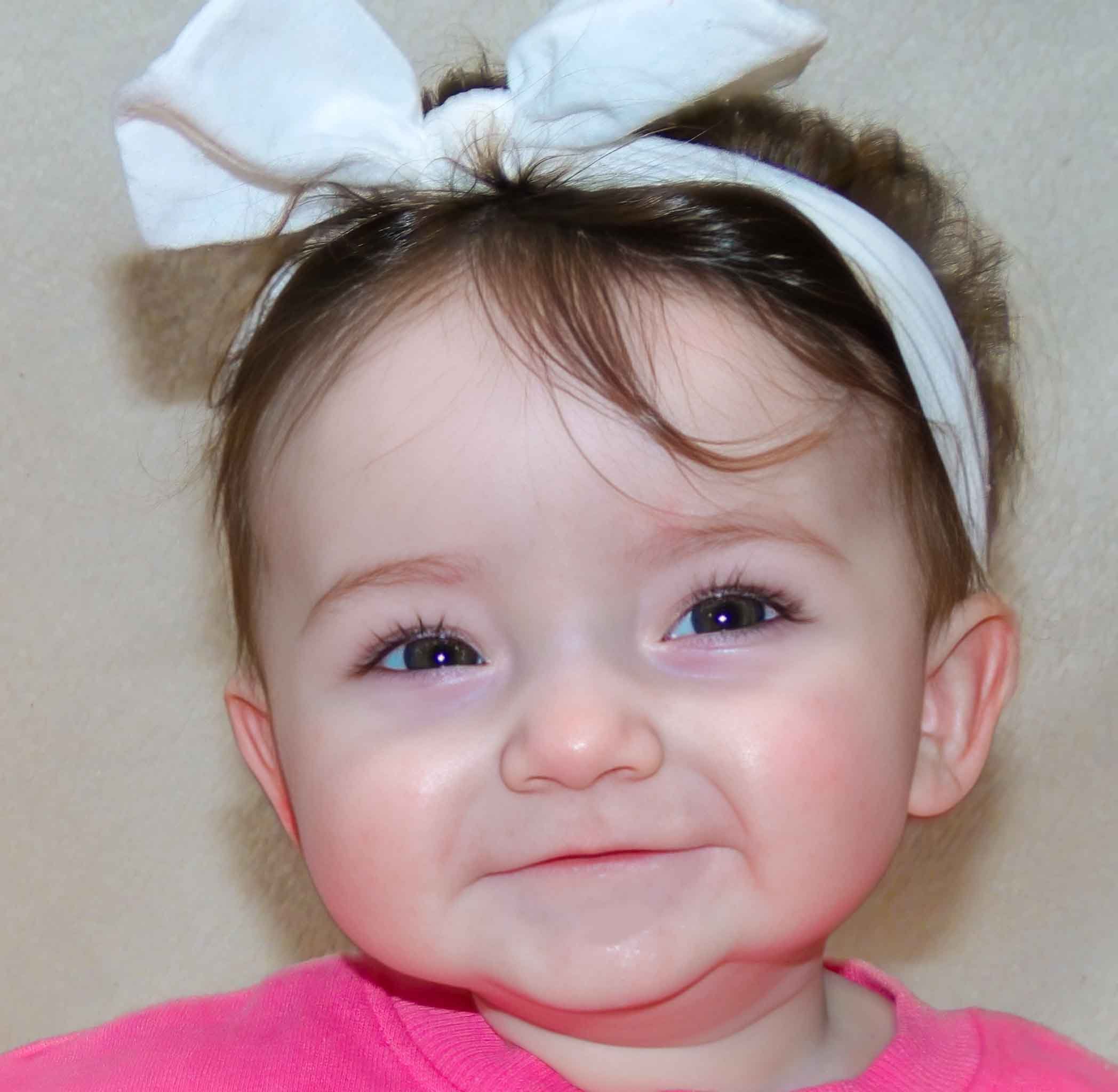 Close-up of a young child's face, showing bright blue eyes, light skin, and brown hair, wearing a pink top and a white headband.