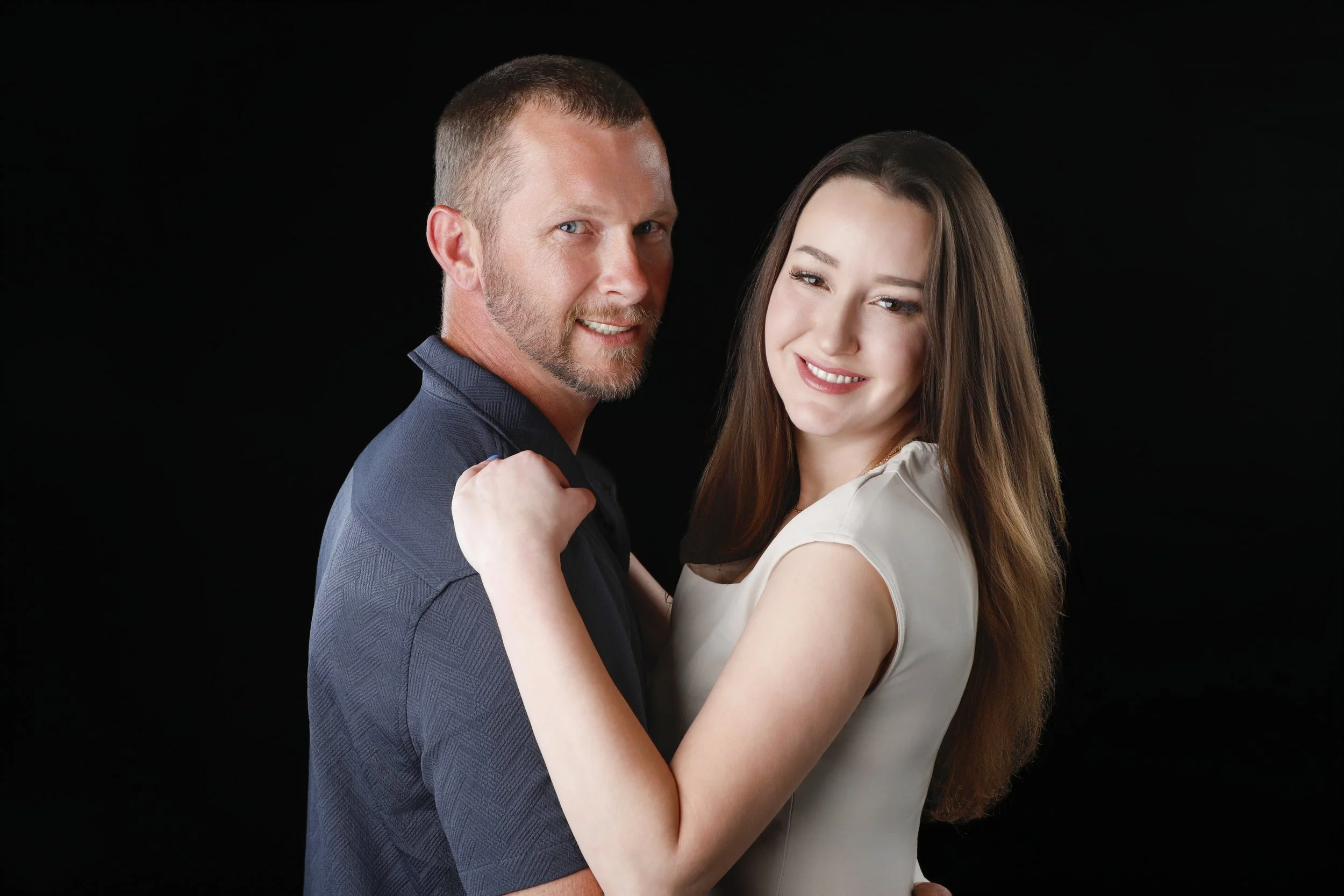 A man and a woman embracing and smiling at the camera against a black background.