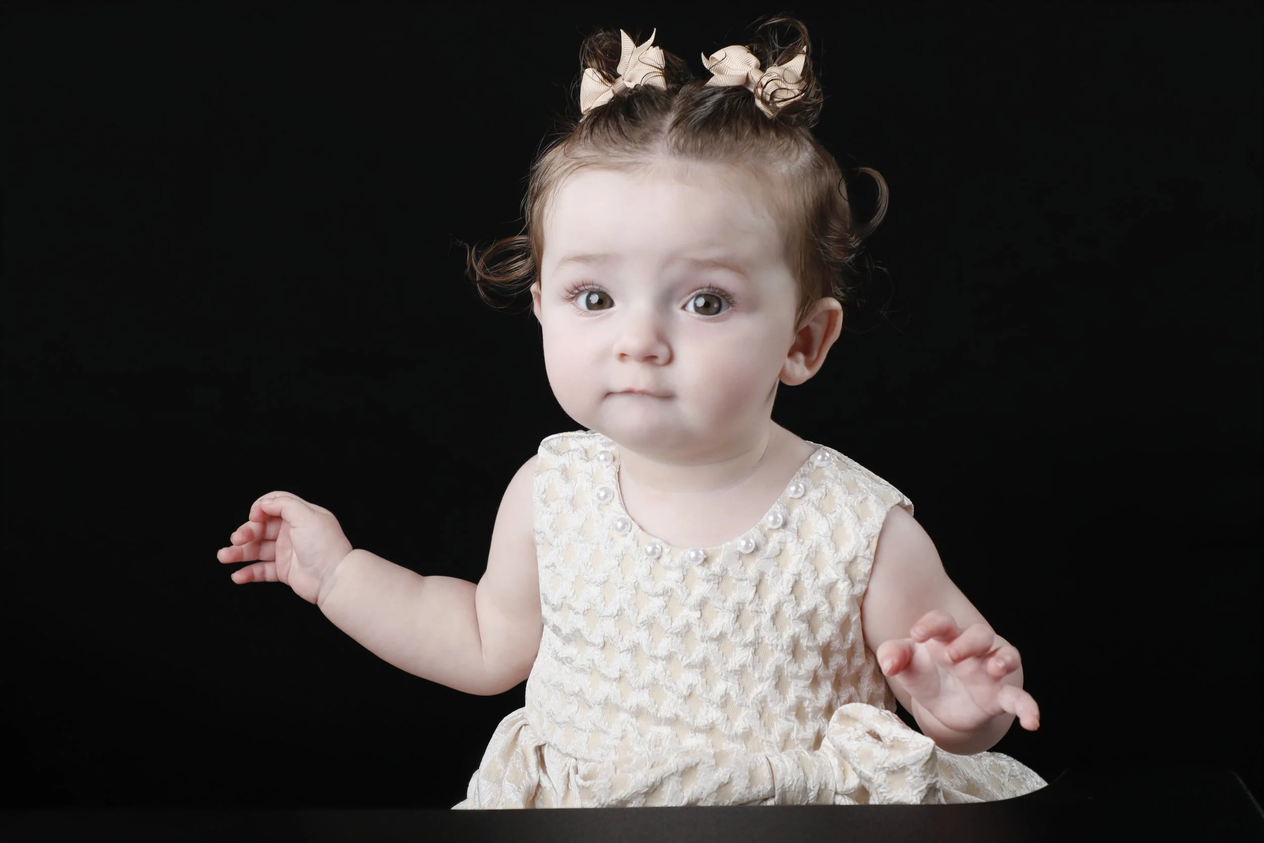 A young girl with brown curly hair styled in pigtails held with beige bows, wearing a cream-colored dress with pearl embellishments, looks curiously at the camera against a black background.