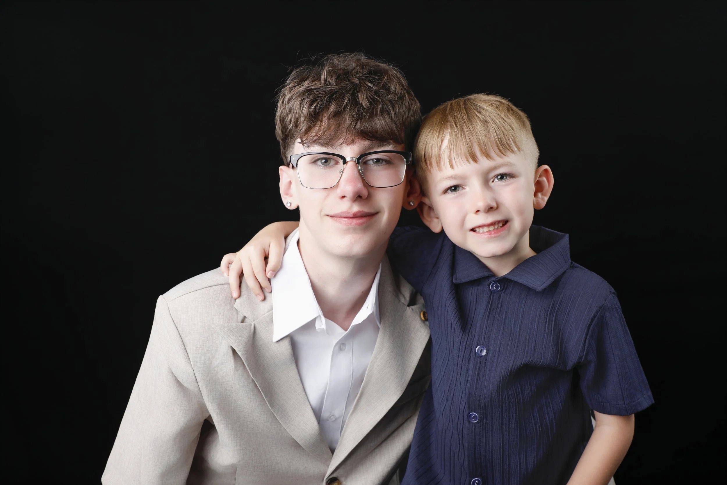 A woman with short brown hair, glasses, and earrings, wearing a light-colored blazer, is sitting next to a young boy with blonde hair, wearing a dark blue shirt. They are smiling and posing together against a black background.