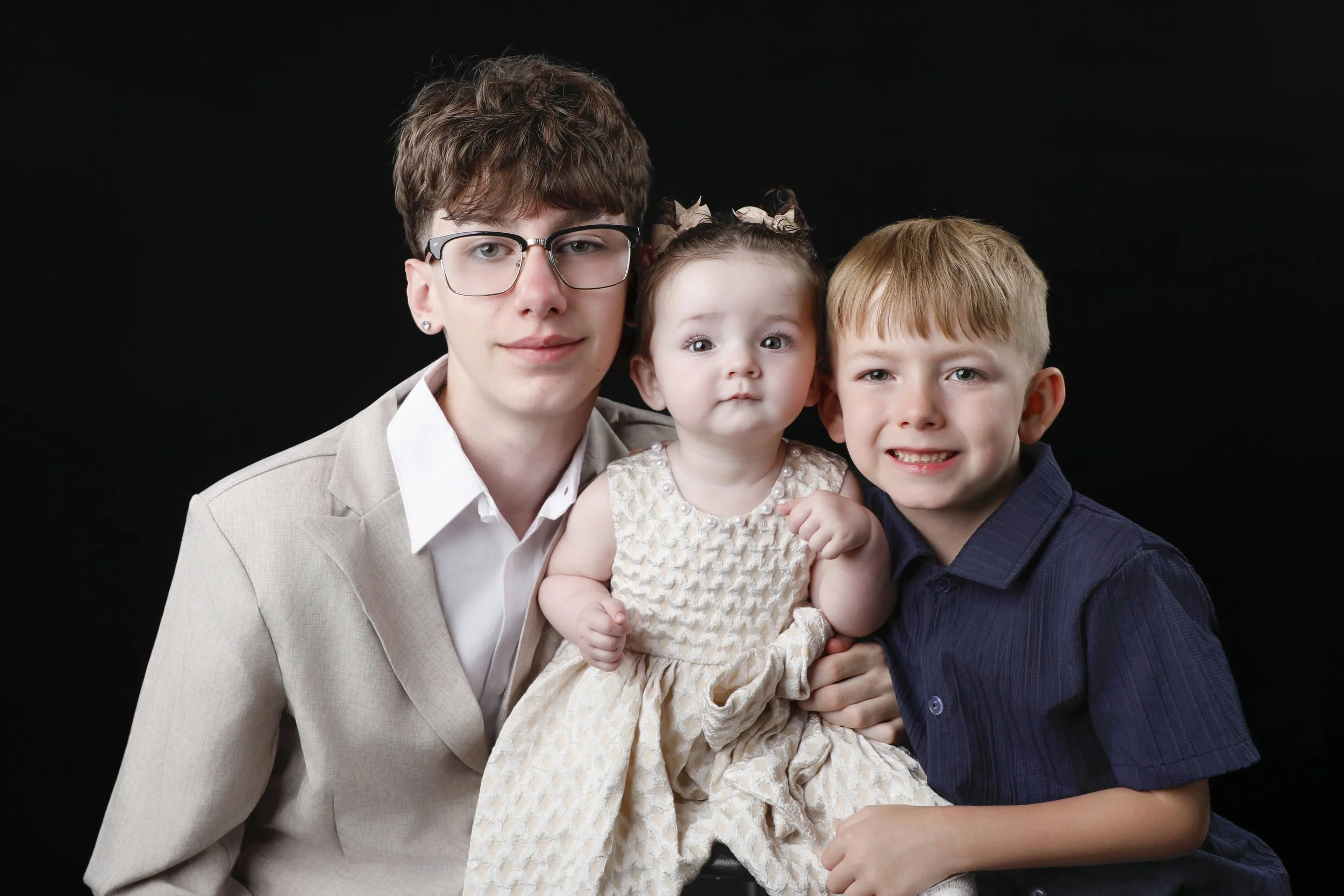 Portrait of a young man with glasses holding a young girl and boy against a black background.