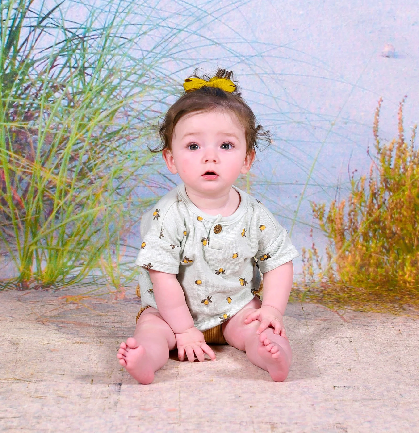 A young child with curly brown hair, wearing a yellow hair bow and a cream-colored shirt with a lemon print, sitting on a wooden floor amid tall grasses, with a pastel-colored background.