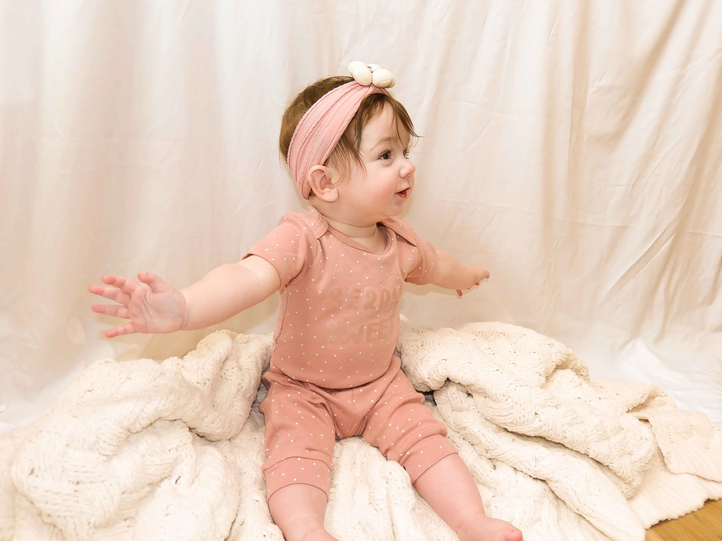 A young girl with short brown hair, wearing a pink headband with a bow and a peach-colored polka dot shirt, smiling and reaching out, sitting on a soft, cream-colored blanket.