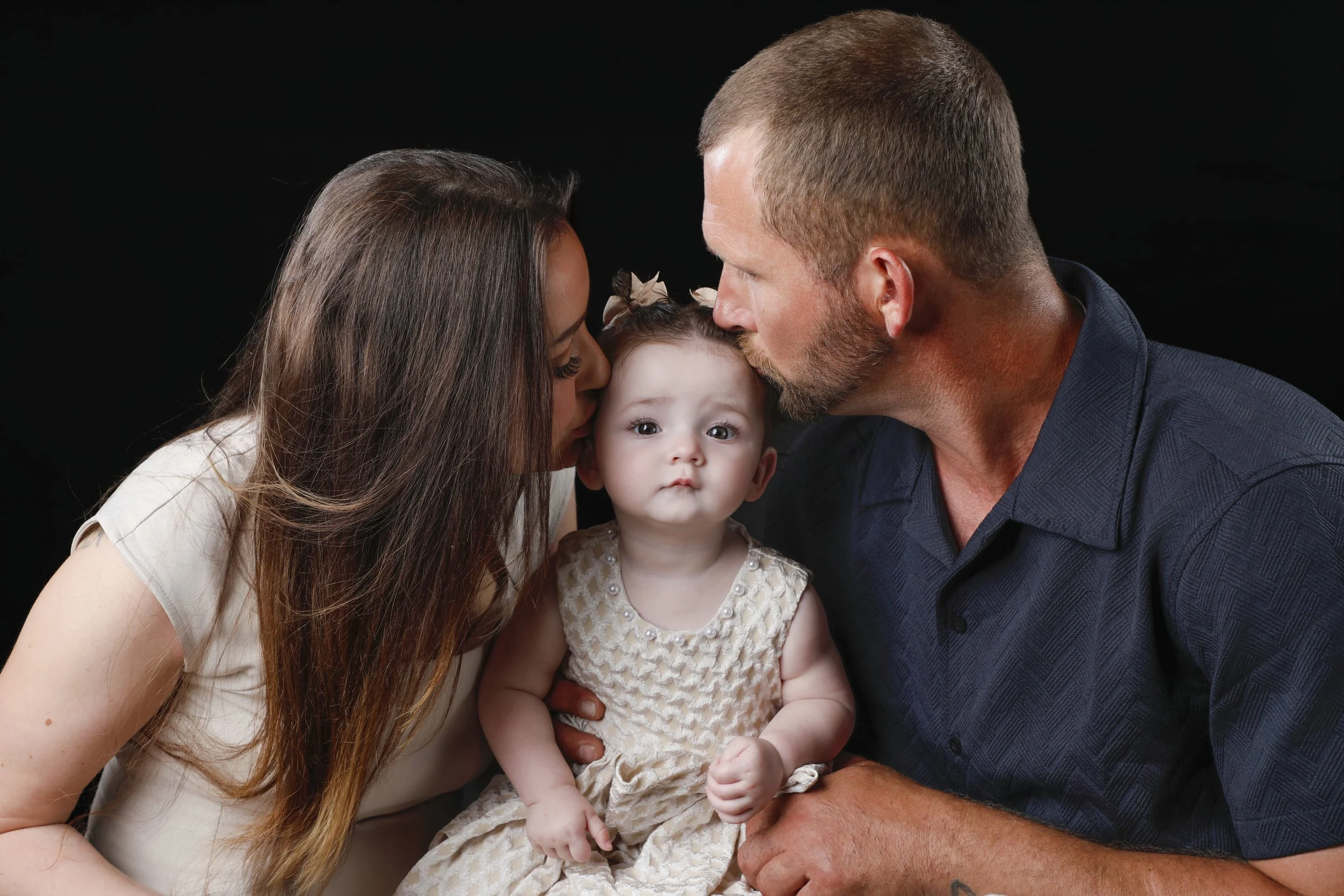A family of three, including a woman, a man, and a young girl, close together against a black background. The woman and man are kissing the girl on each cheek.