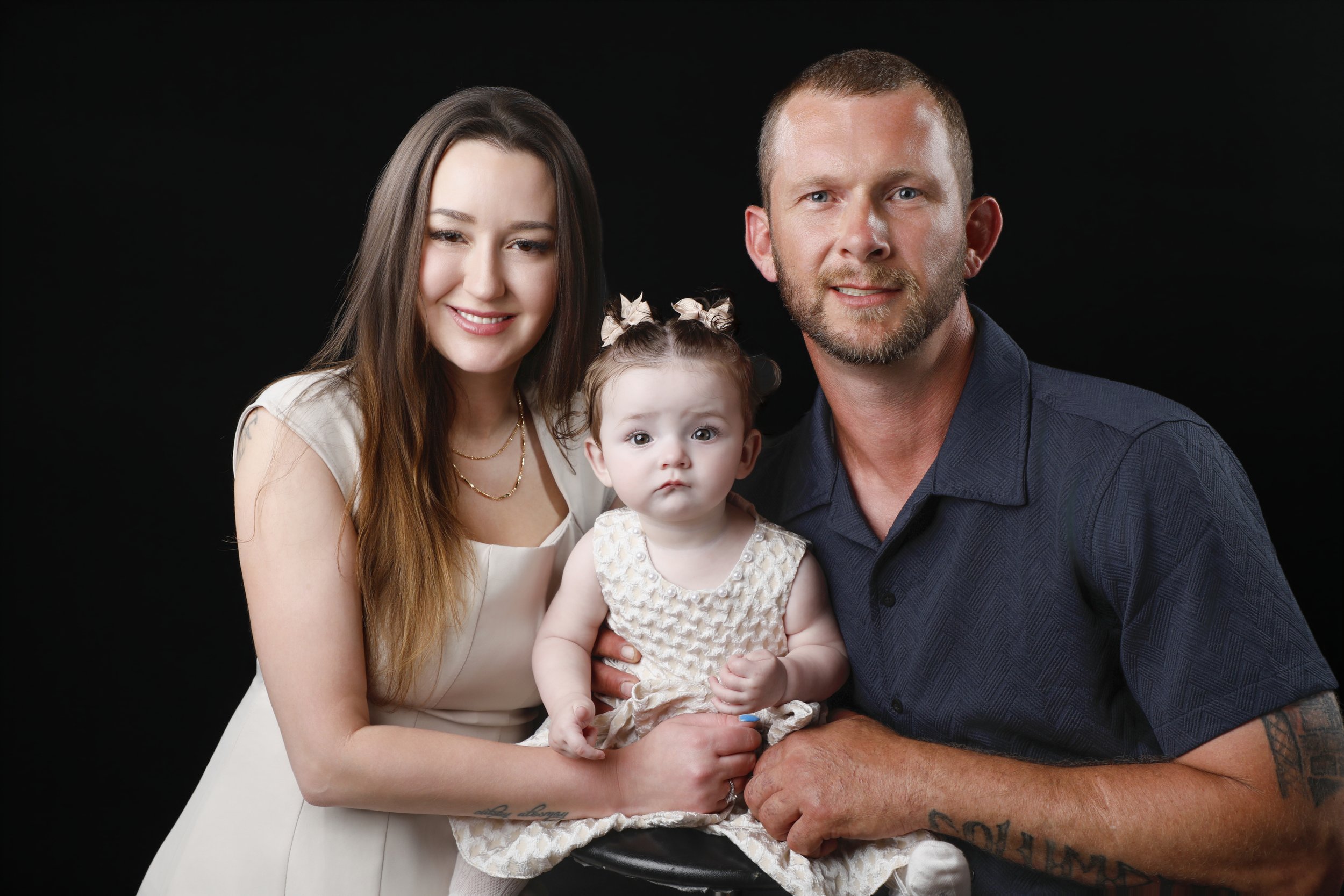 A family of three sitting together against a black background. The mother has long brown hair and is smiling, wearing a white dress. The father has short hair and a beard, wearing a blue shirt, and is smiling. The young daughter has curly hair tied w