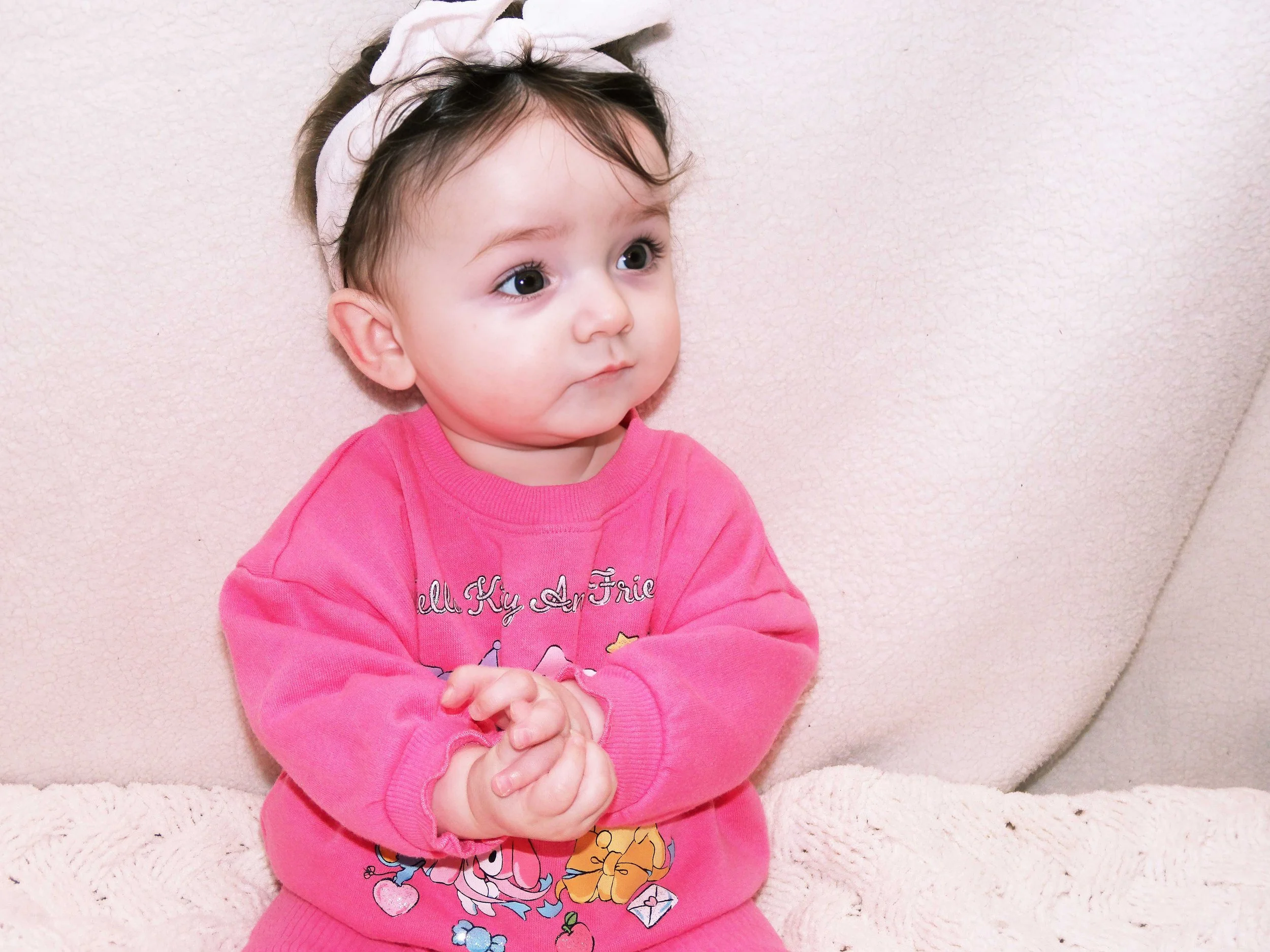 A young girl wearing a pink shirt with cartoon characters and a white headband with a bow, sitting against a light-colored wall with her hands clasped.